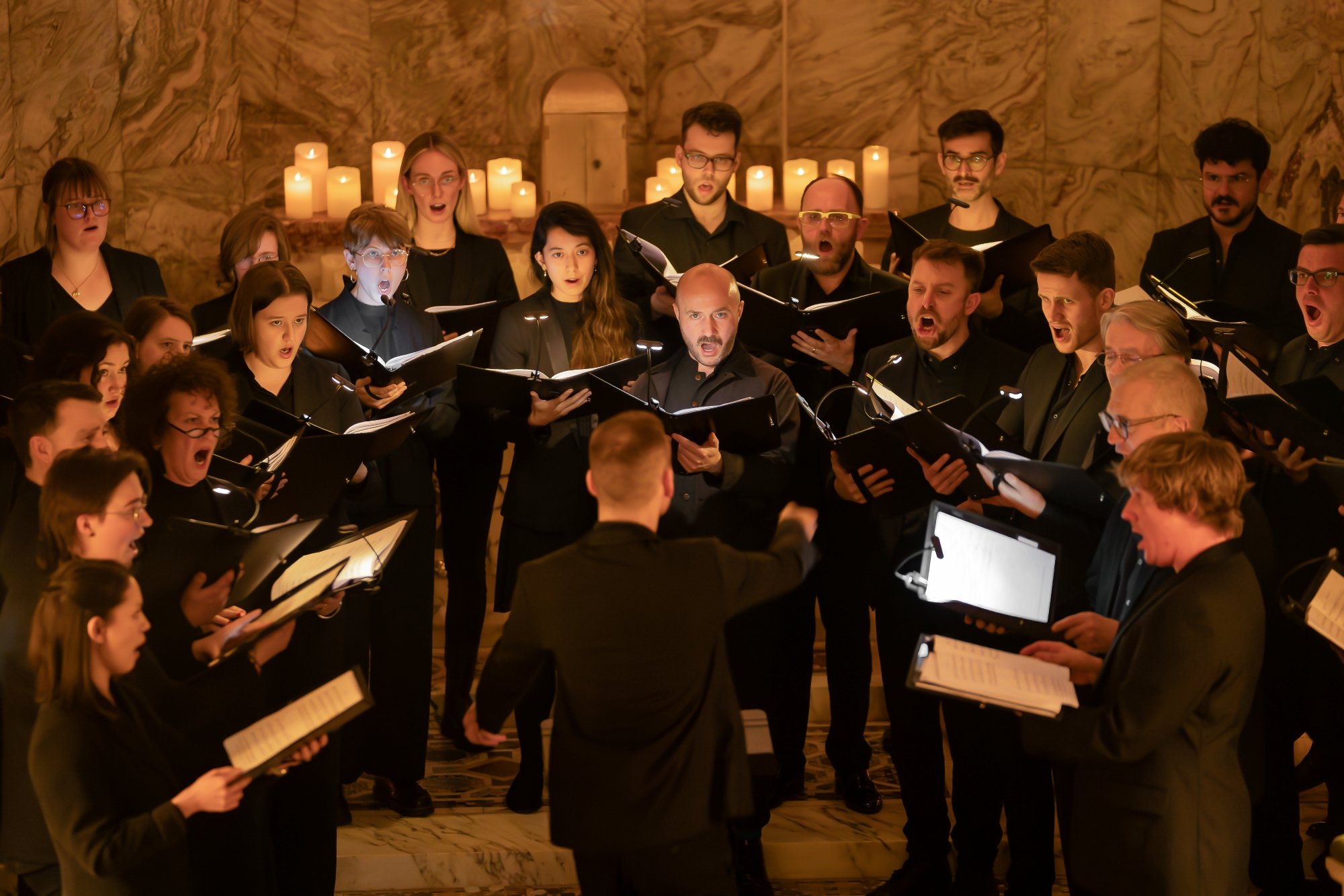 A choir performing in a church with candles in the background, led by a conductor.