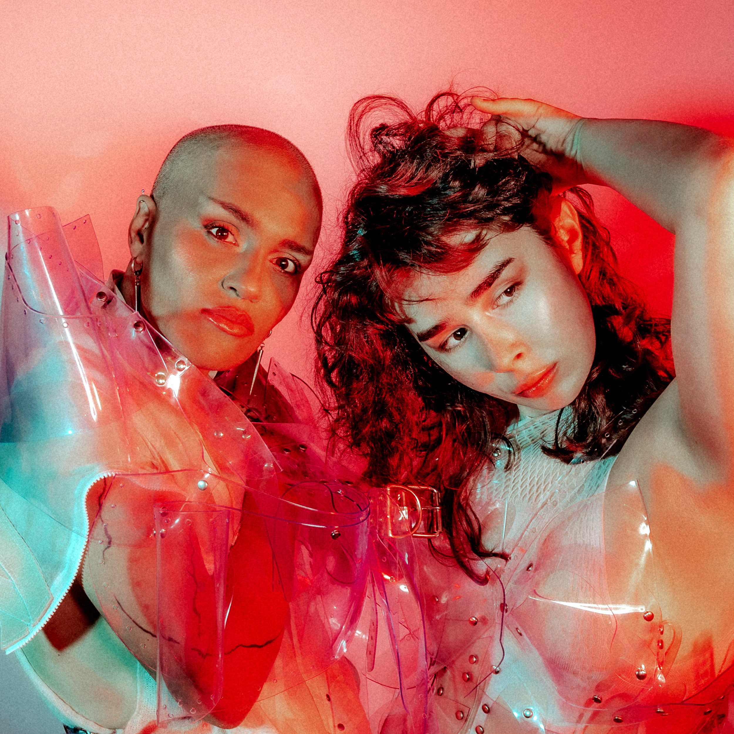 Two women posing together in colorful, shiny, avant-garde outfits with a pinkish-red background.