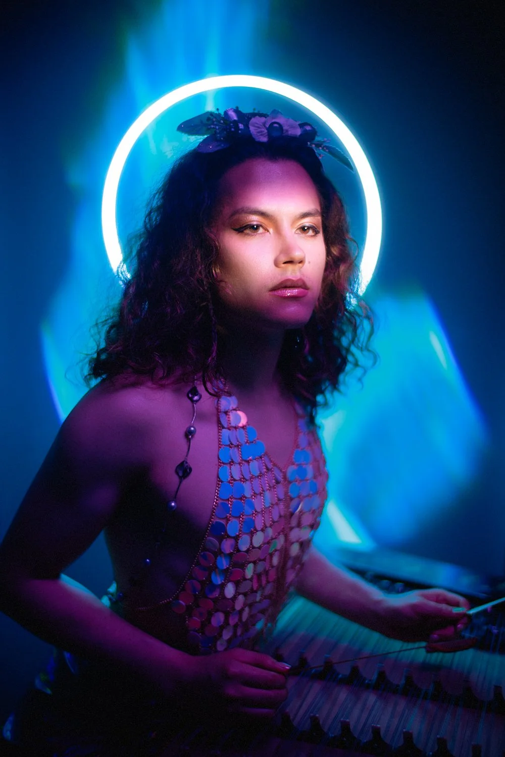 A woman with curly hair wearing a flower headband and jewelry, illuminated by neon lights, appears to be playing a xylophone.