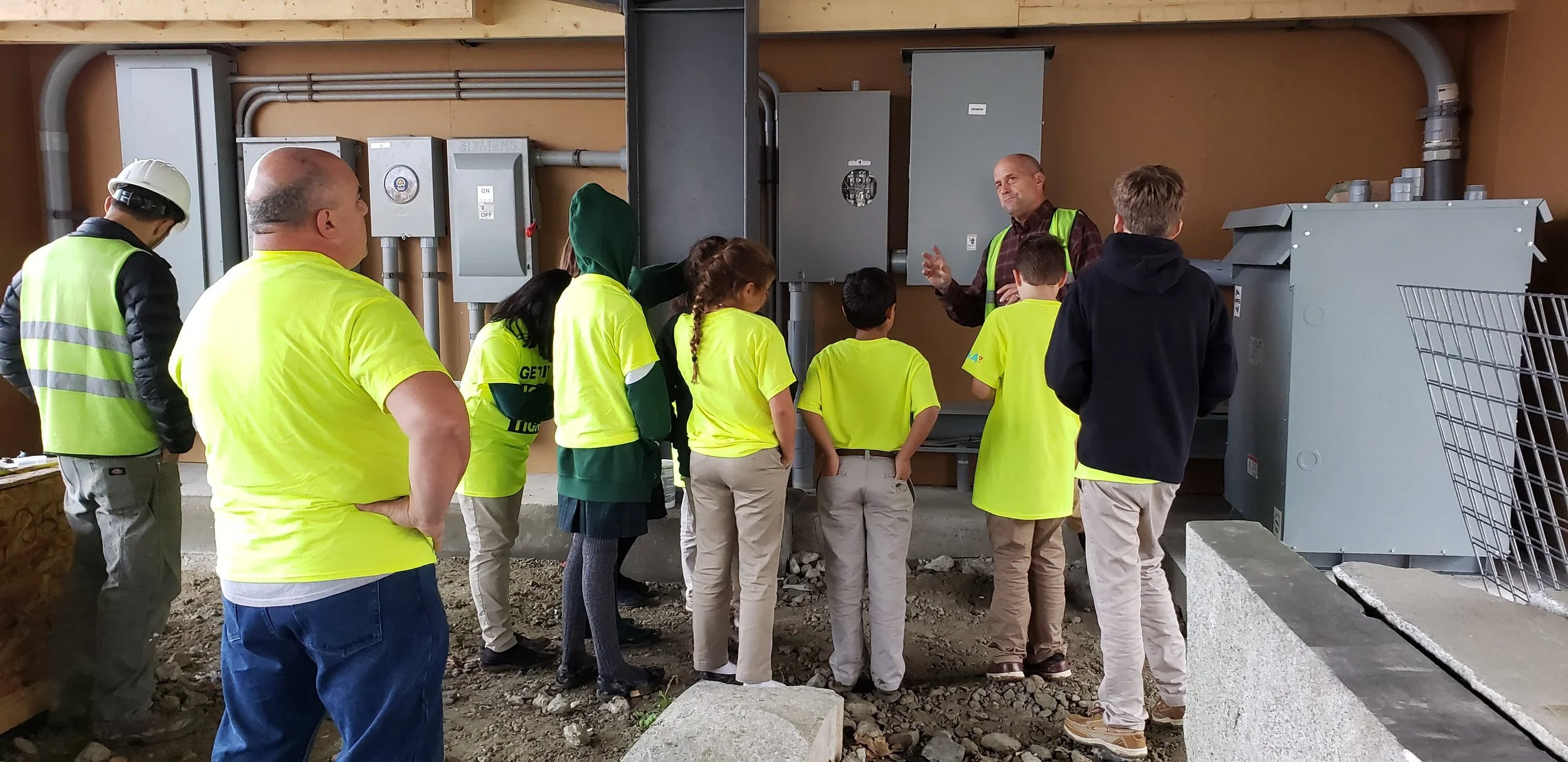 Group of children and adults in bright yellow safety vests and casual clothing listening to a man explaining electrical equipment in an industrial setting.