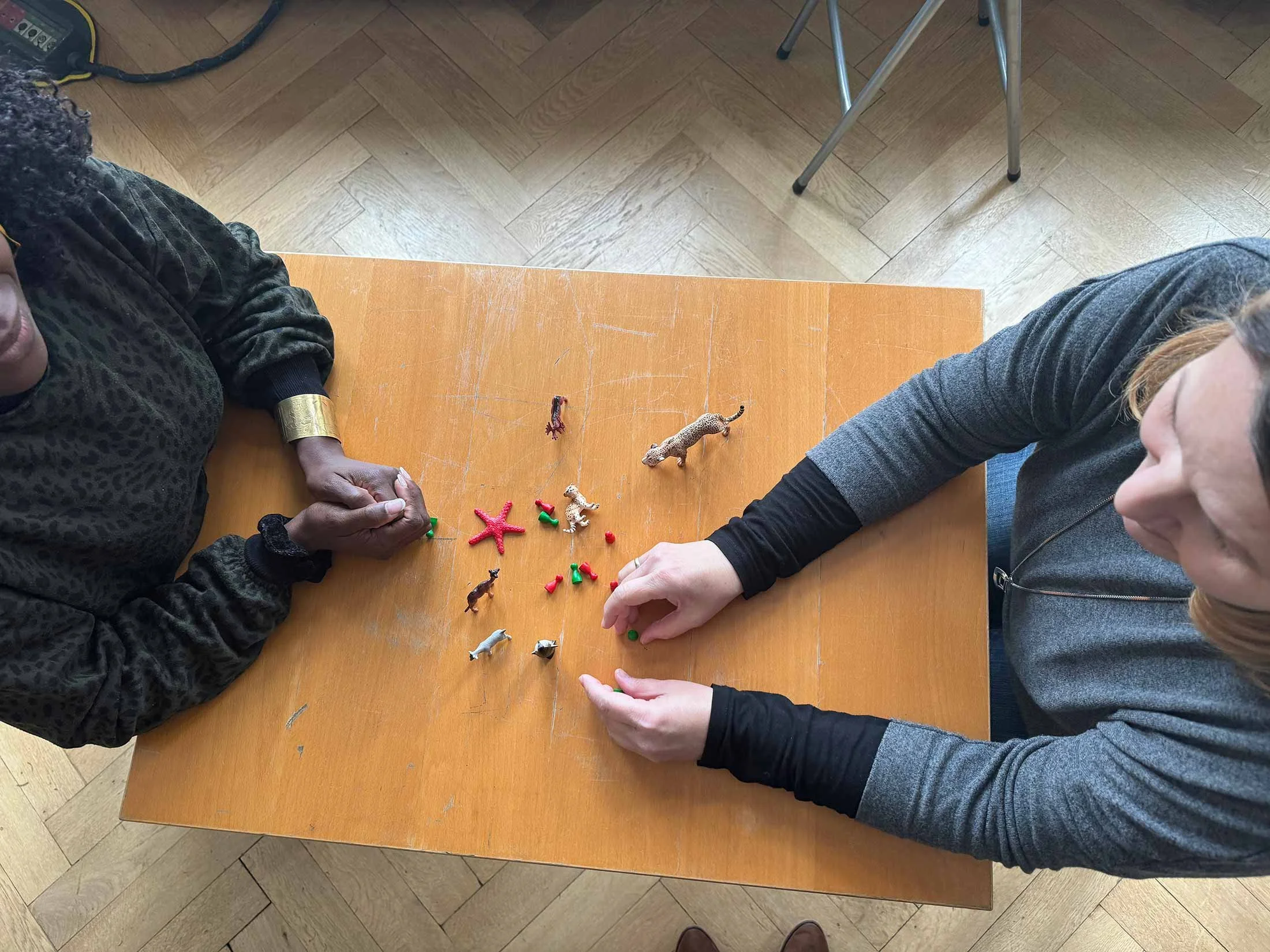 Two people sitting at a wooden table with small toy animals and objects, including a starfish and plastic animals, seen from above.