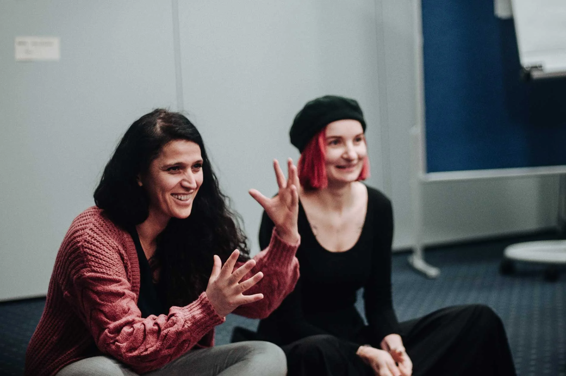 Two women seated in a classroom or meeting room, smiling and engaging in conversation, with one woman gesturing with her hand.