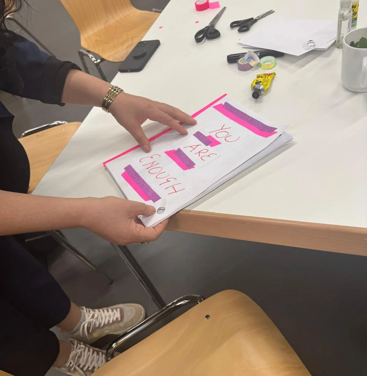 A person holding a notebook with the phrase "You are enough" written on it, highlighted with pink and purple tape, in a workspace with scissors, tape, a phone, and a mug on the table.