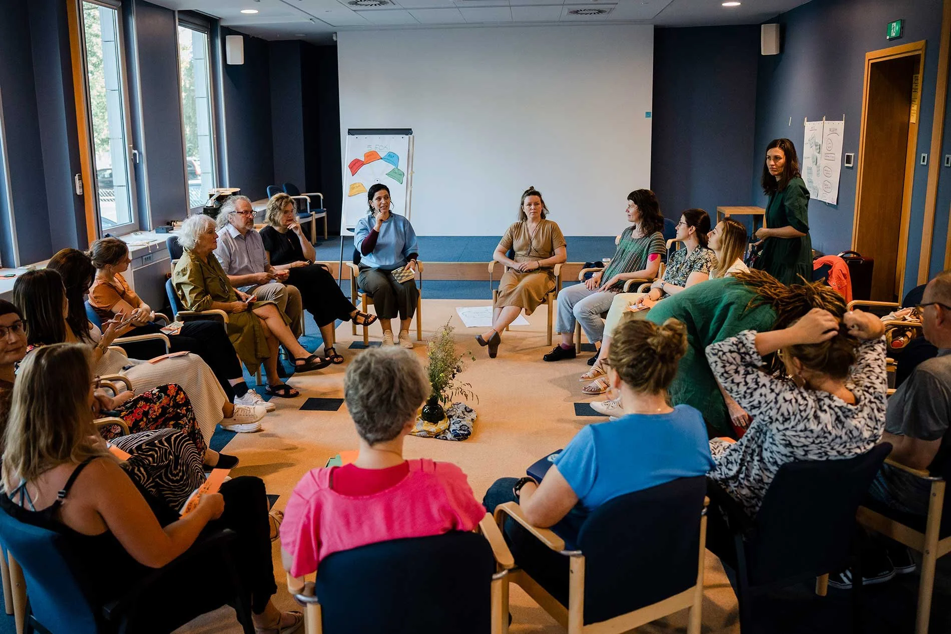 A diverse group of people participating in a workshop or discussion in a conference room. Some are seated around a semi-circle, while one woman stands and speaks. The room has large windows, a whiteboard with colorful charts, and posters on the walls