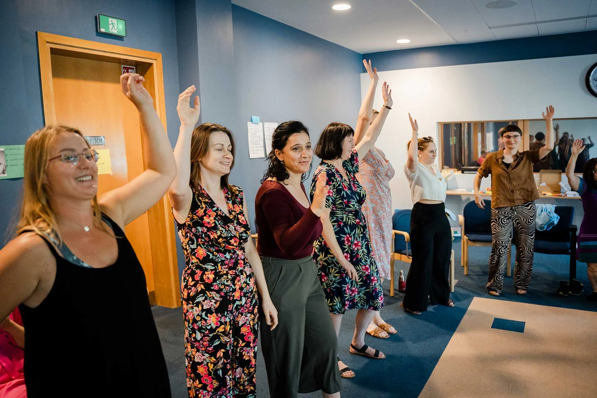 A group of women standing in a row with their hands raised, participating in an activity inside a room with gray walls and a ceiling with recessed lighting. Some women are smiling and looking forward. The setting appears to be a workshop or meeting.