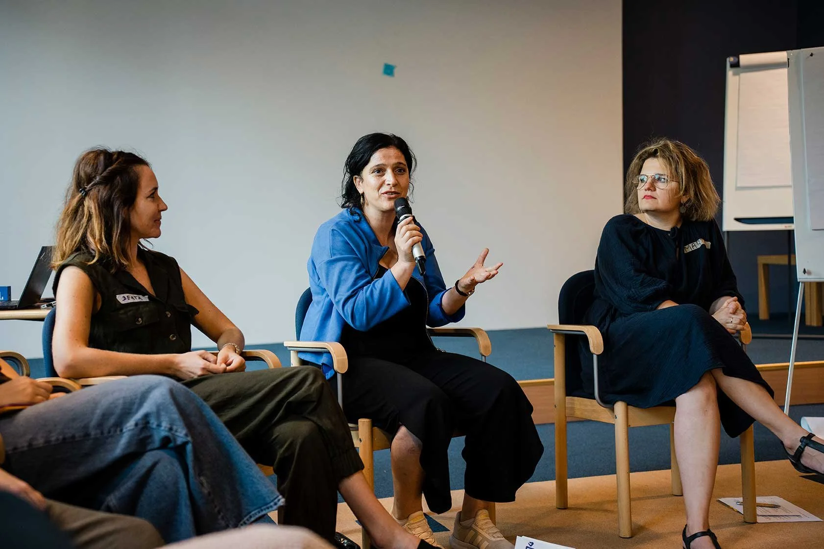 Three women seated in a panel discussion, with the woman in the middle speaking into a microphone, in a professional setting with a whiteboard on the right.