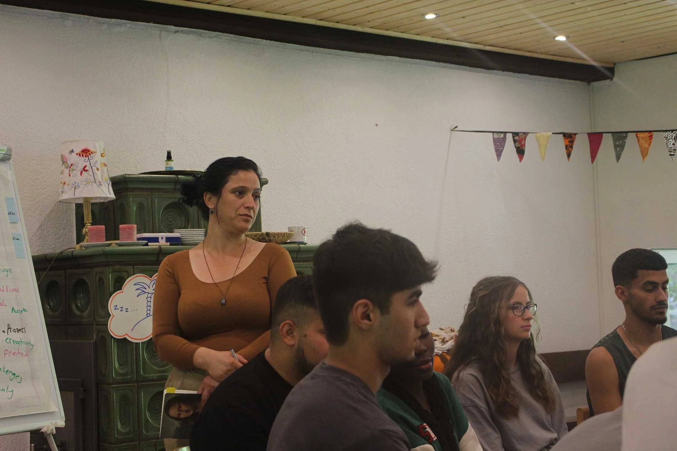 A woman with dark hair in a bun and wearing a brown top stands among seated young adults in a room decorated with colorful bunting. She appears to be attentive or listening, with a serious expression. In the background, there is a whiteboard, a lamp 