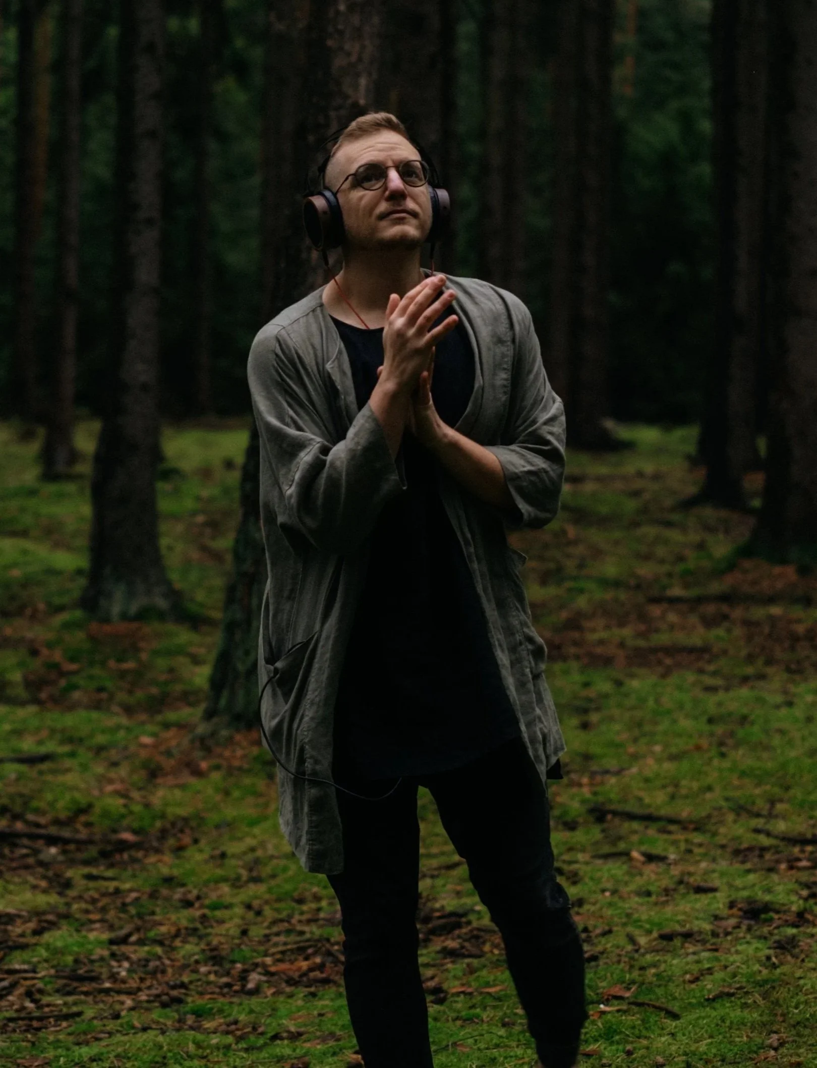 A man stands in a dense forest during twilight, wearing headphones and glasses. He has short hair and wears a gray jacket over a black shirt, with his hands clasped together near his chest.
