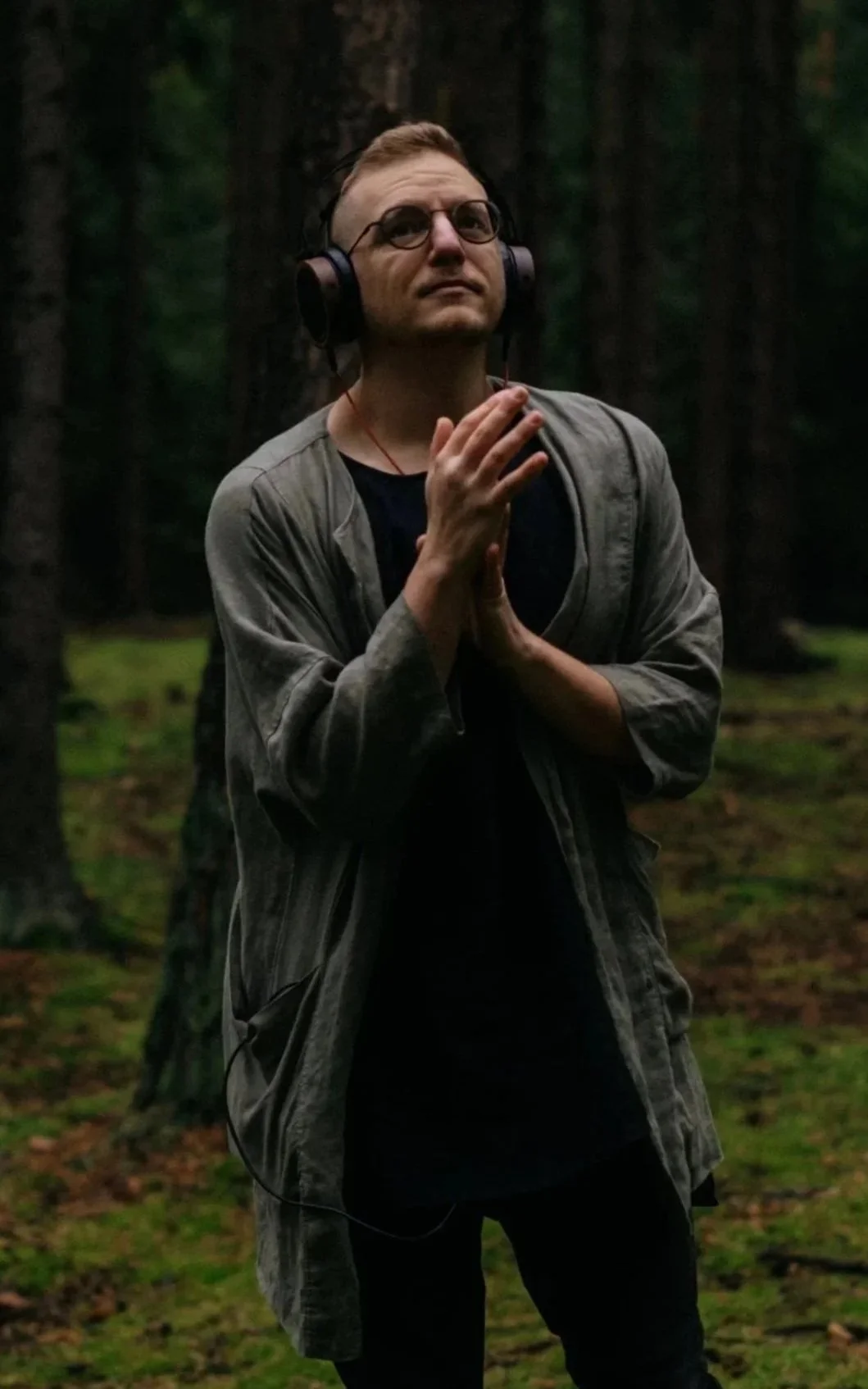 A man with glasses and headphones standing in a forest, listening to music with his hands clasped together.