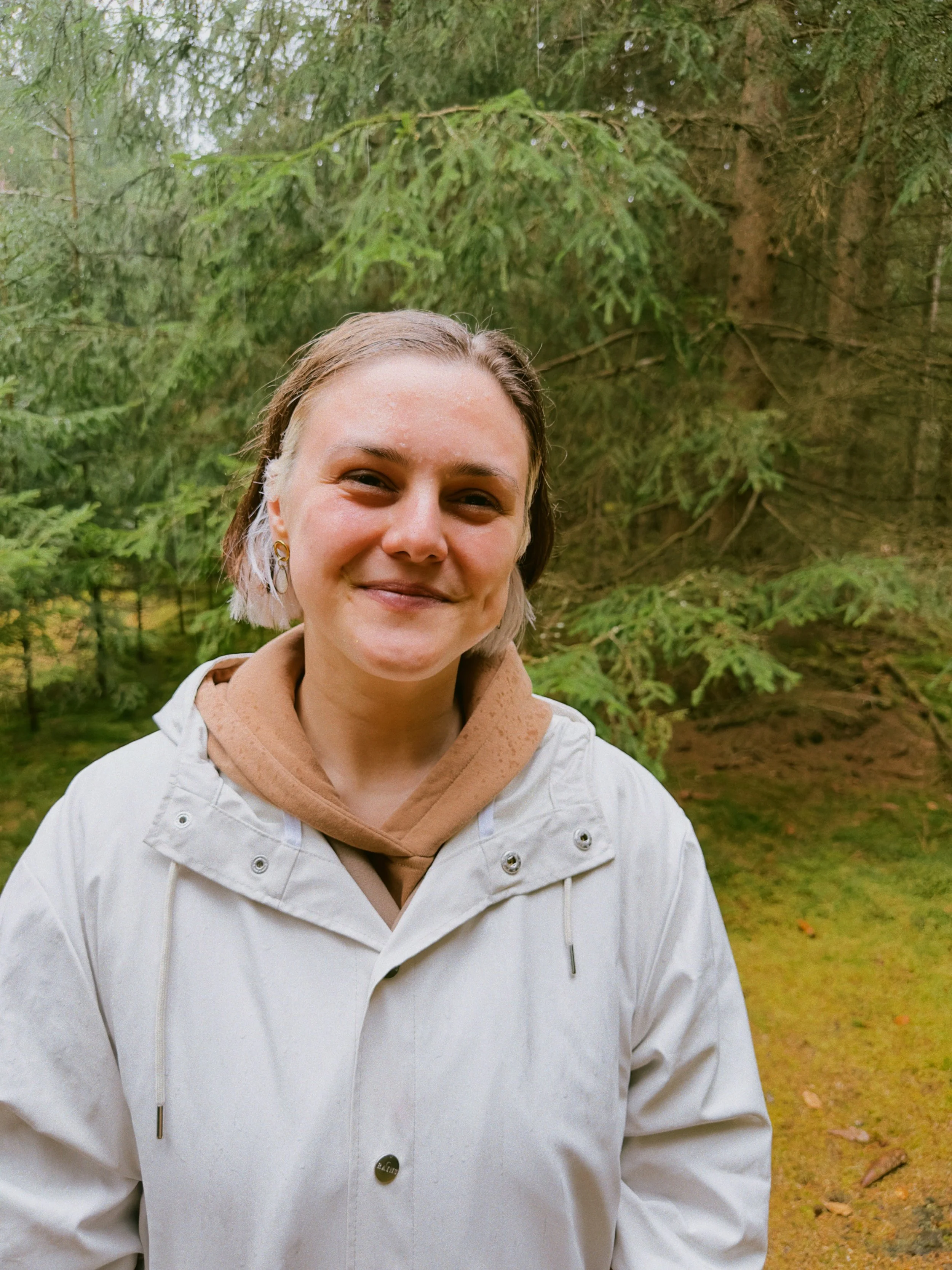 A woman smiling outdoors in front of green trees, wearing a white jacket and a tan hoodie, standing in the rain.