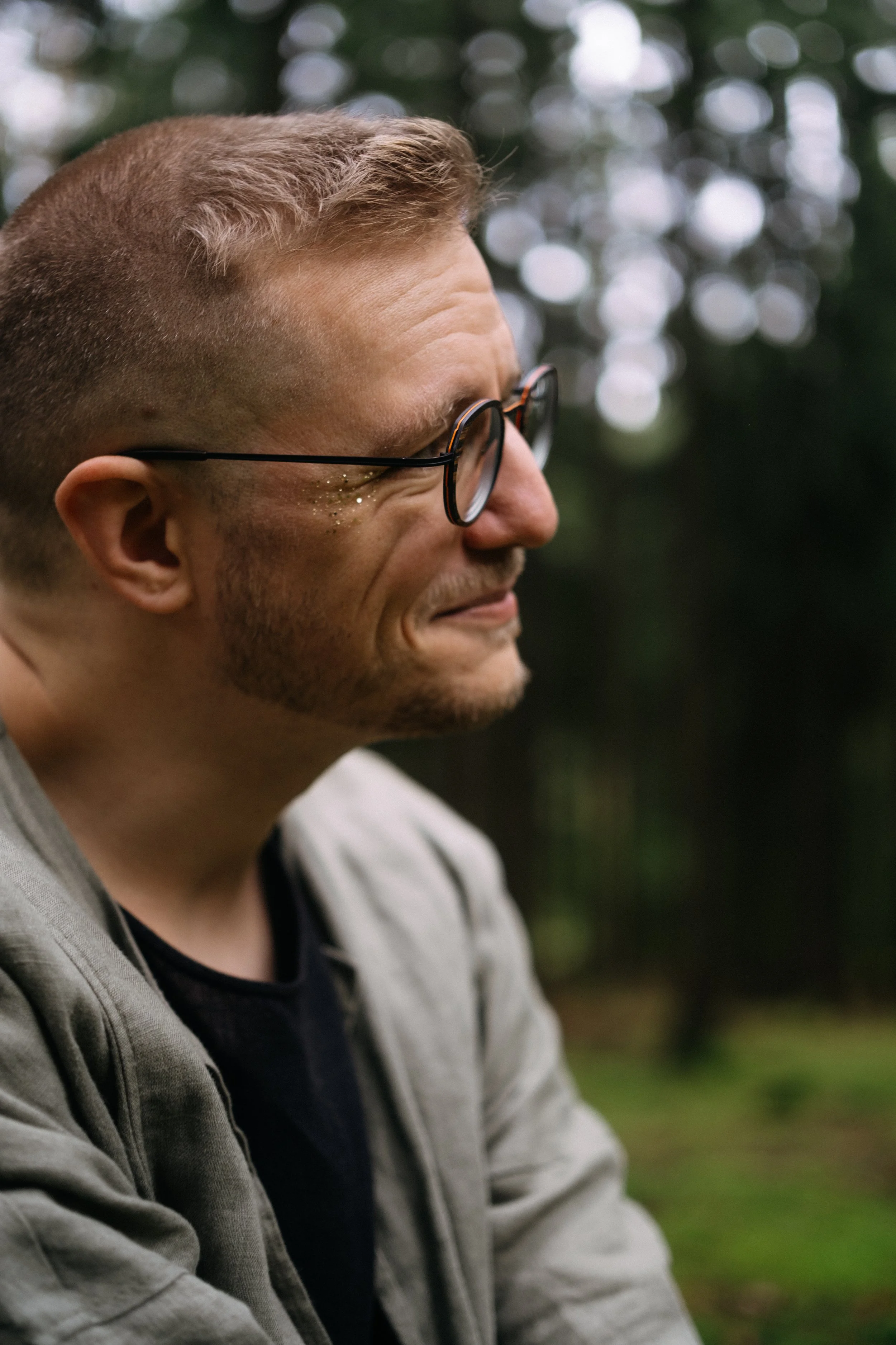 A man with short, light brown hair and glasses, smiling with his eyes closed outdoors, with blurred green foliage in the background.