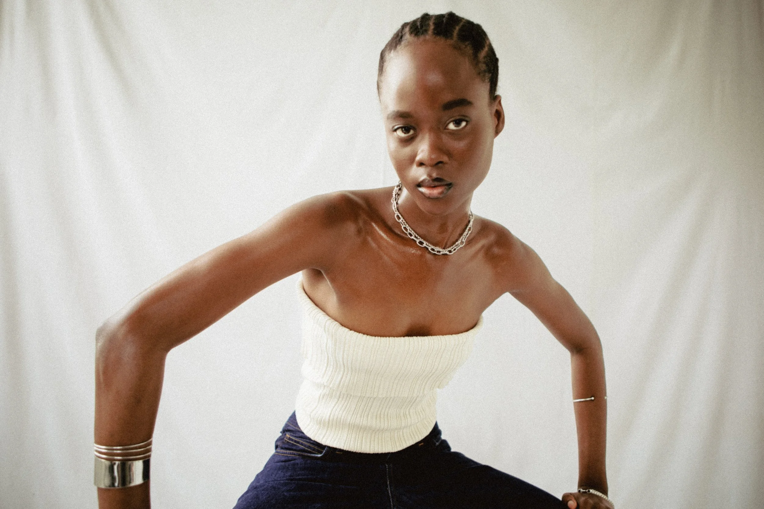 Jeune femme en top blanc et jeans foncés, portant des bijoux en argent, posant de face devant un fond blanc.