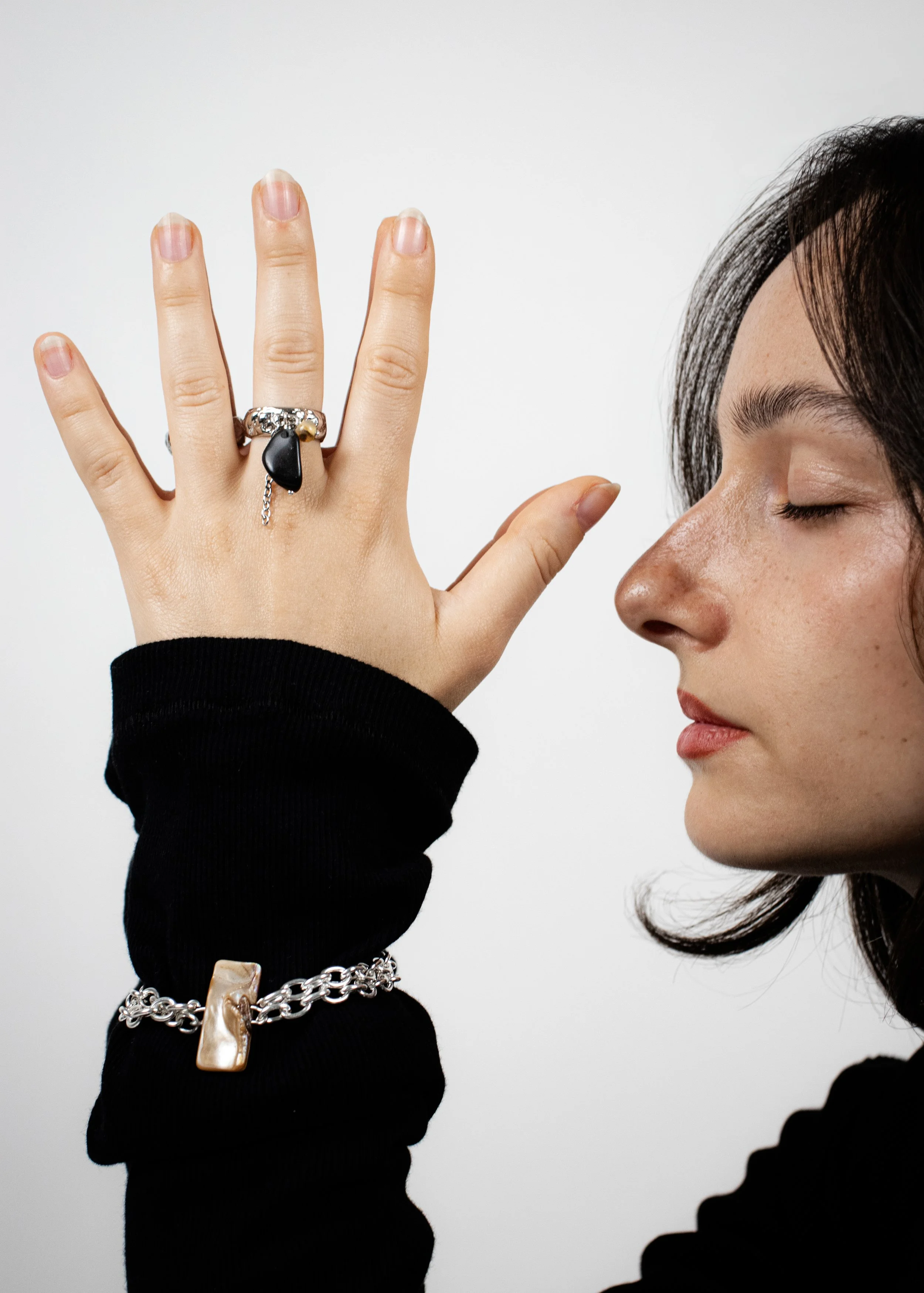 Une femme avec des yeux fermés montre sa main portant plusieurs bijoux, dont une bague avec une pierre noire et une chaîne avec un pendentif en or, un bracelet en or avec une pierre semblable, contre un fond blanc.