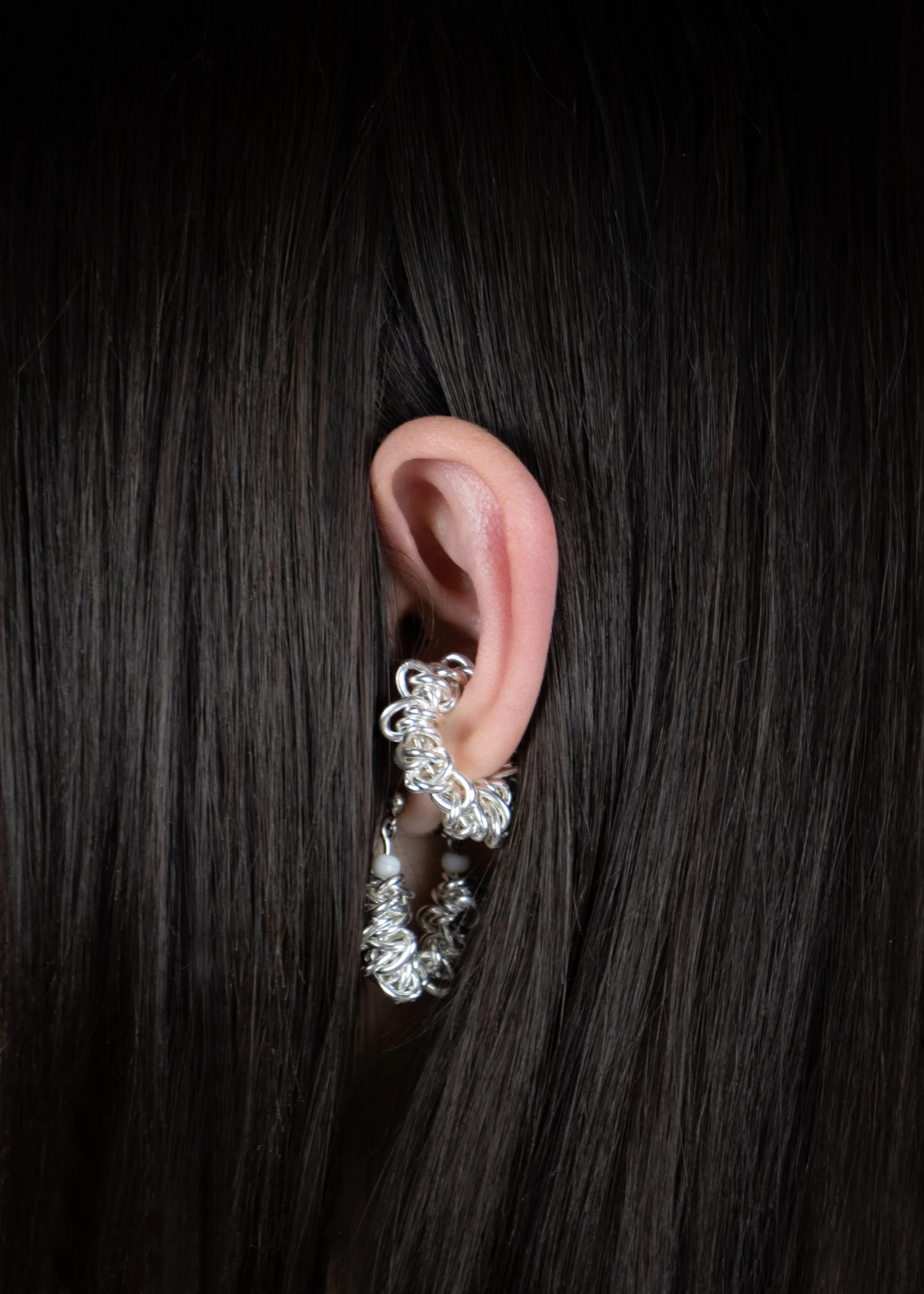 Une oreille porte une boucle d'oreille en formes de chaînes en métal argenté avec des perles blanches, entourée de cheveux noirs lisses.