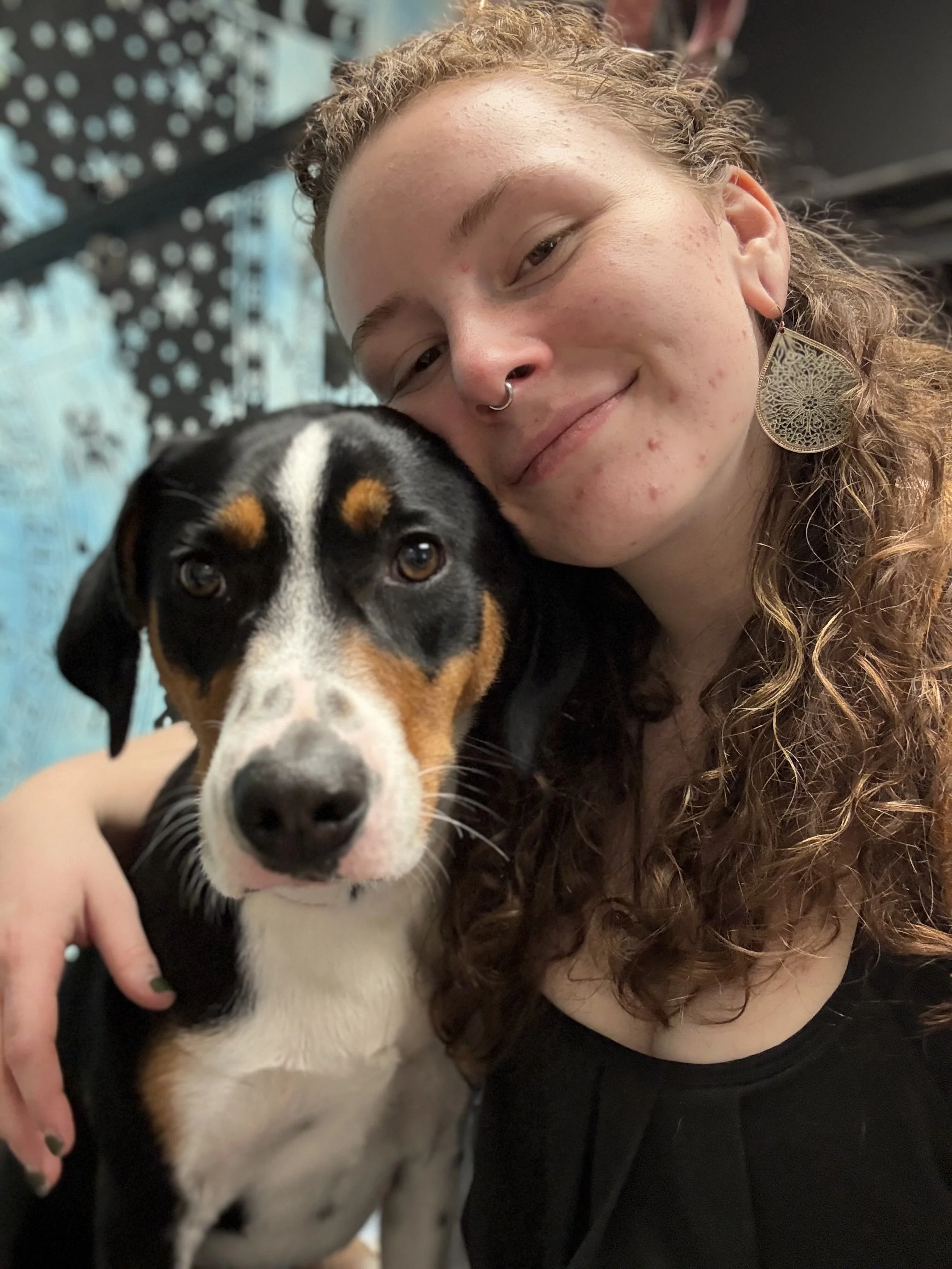 A person with curly hair and light skin, wearing large earrings and a septum piercing, is smiling and holding a black, white, and tan dog close to their face. The background features a blue and black patterned wall with snowflake designs.