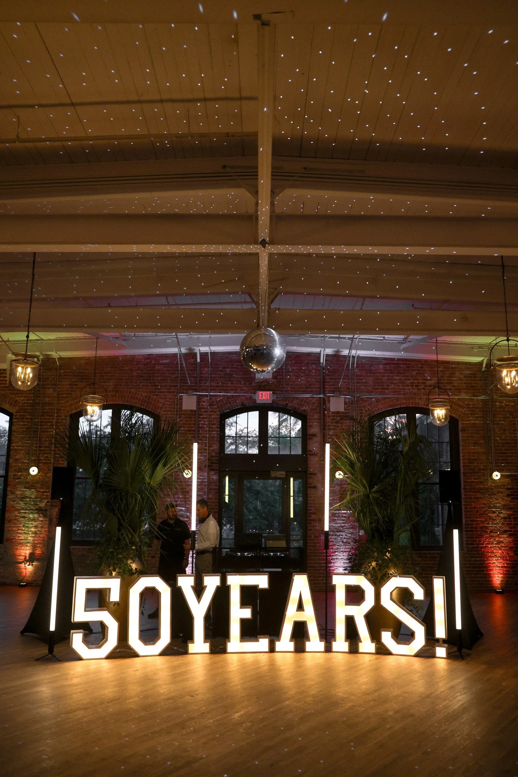 Celebration event with illuminated sign reading '50 YEARS!', large disco ball, palm plants, hanging lights, brick walls, and a DJ setup with two men talking inside a decorated venue.