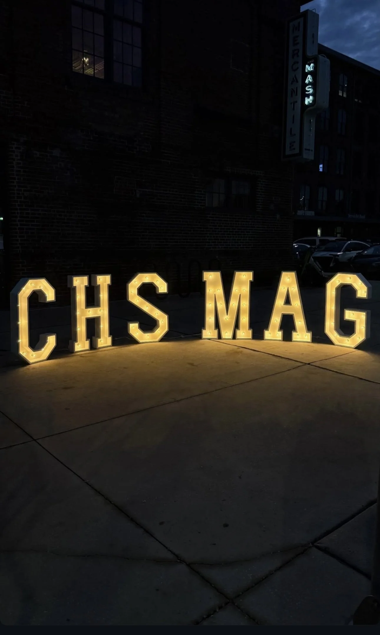Illuminated marquee letters spelling 'CHSMAG' on a sidewalk at night, with a brick building and a lit sign reading 'MERCIAL' and 'MASH' in the background.