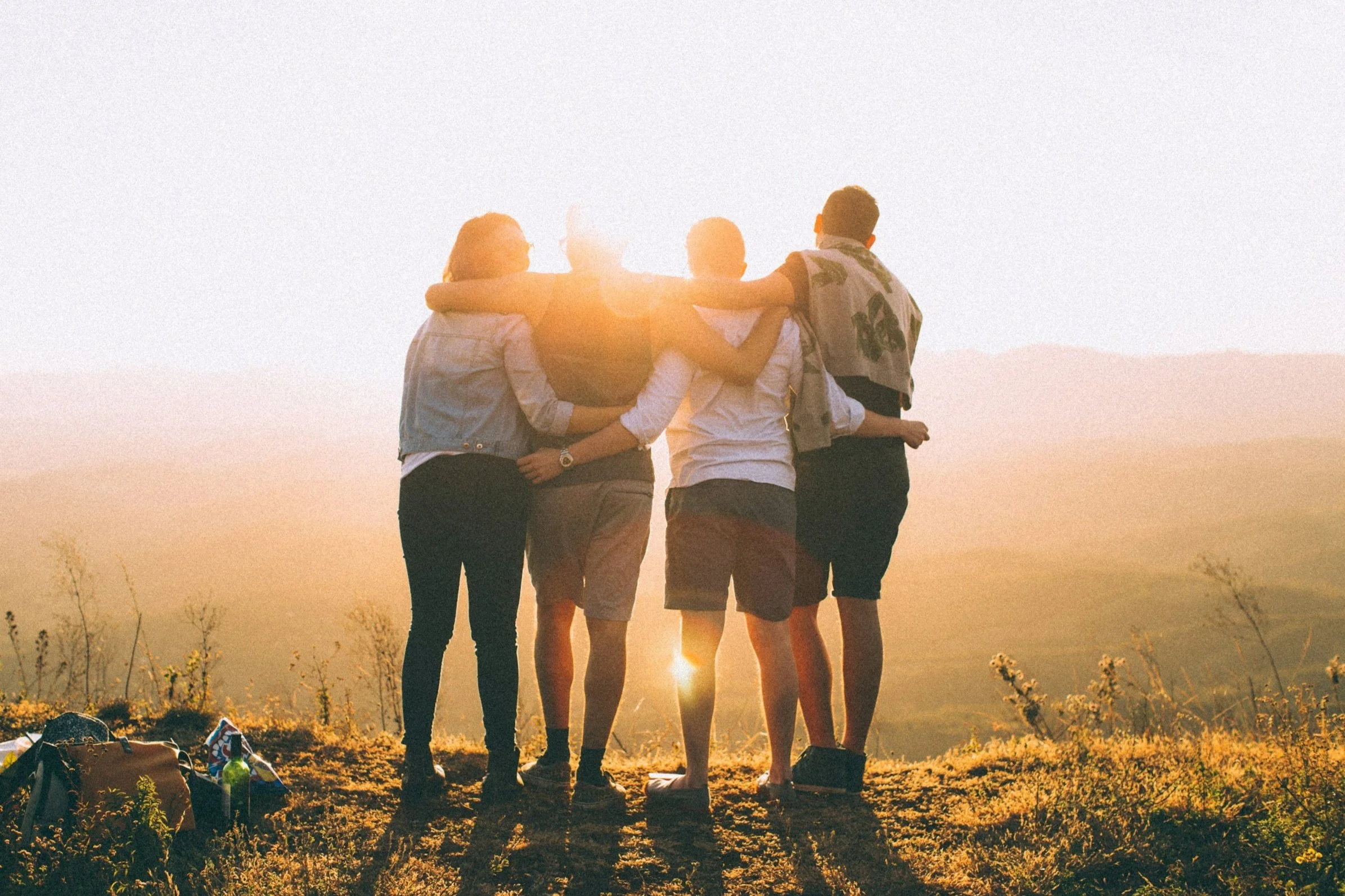Five friends standing together outdoors at sunset, embracing each other with arms around one another on a grassy hill, with backpacks on the ground nearby.