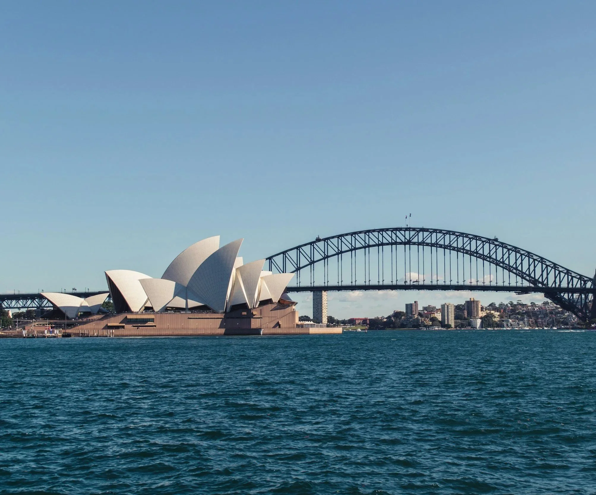 The Sydney Opera House with its distinctive sail-like design on the harbor, and the Sydney Harbour Bridge arching over the city skyline, under a clear blue sky.