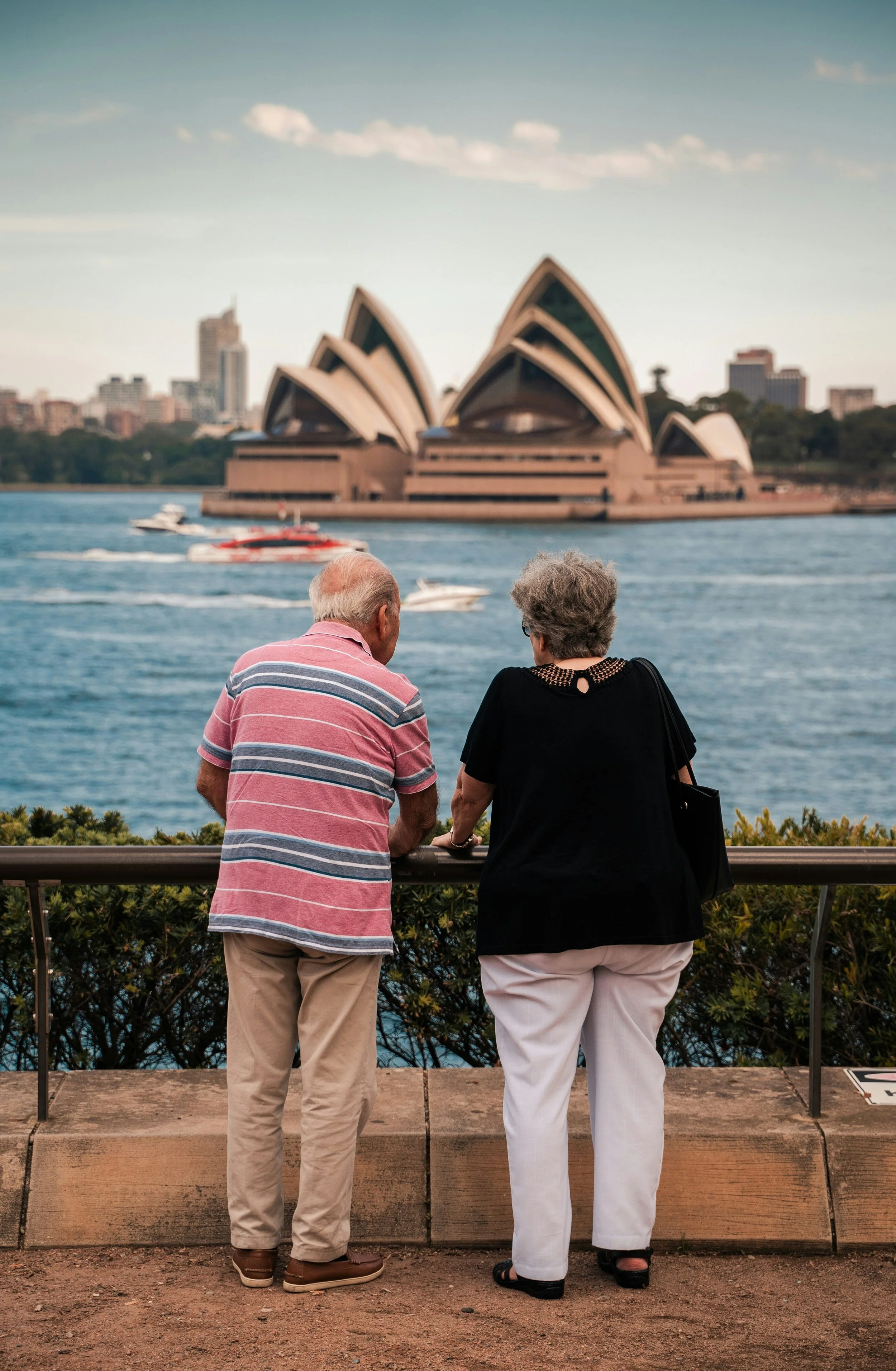 An elderly couple stands by a railing, looking towards the Sydney Opera House across the water in Sydney, Australia.