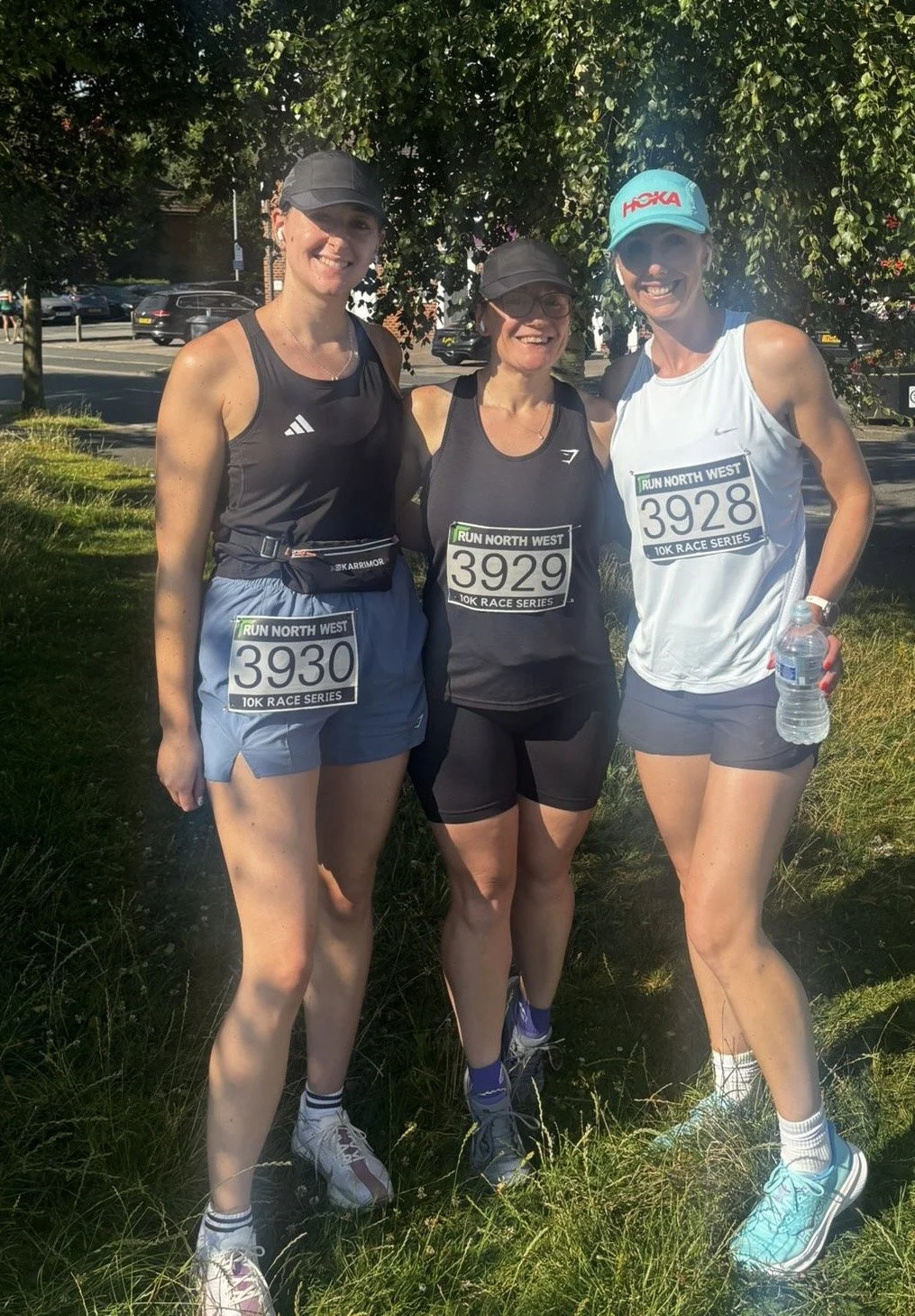 Three women in running gear standing outdoors on grass, smiling, after participating in a race, with race bibs and a background of trees and a street.