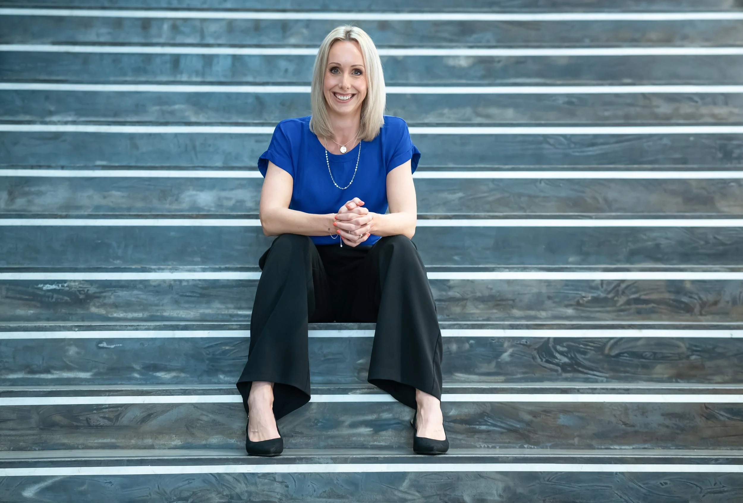 A woman with blonde hair sitting on gray marble stairs, smiling, wearing a blue blouse, black trousers, and black heels.