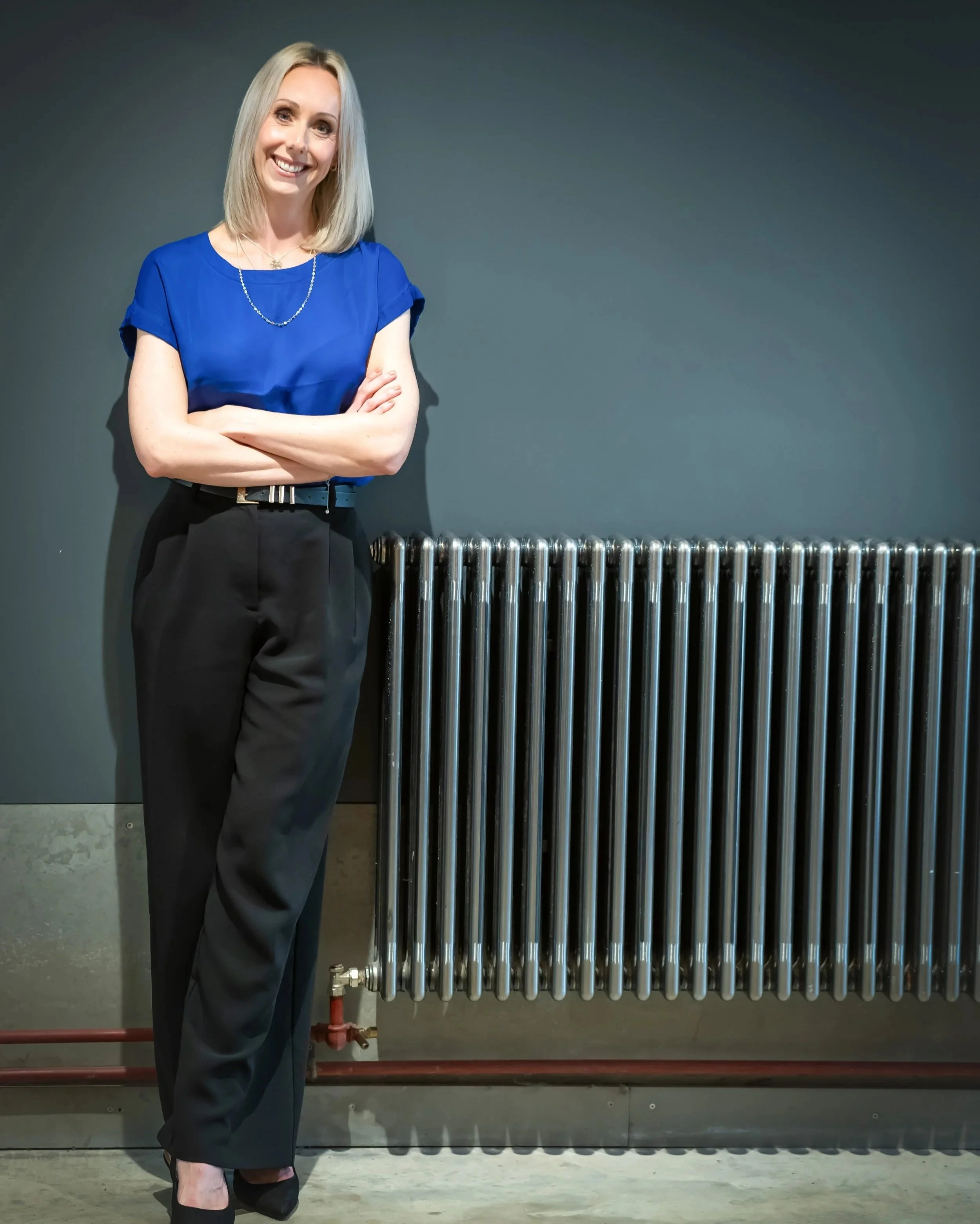 A woman with blonde hair wearing a blue blouse and black trousers, standing with arms crossed next to a metallic radiator against a dark grey wall.