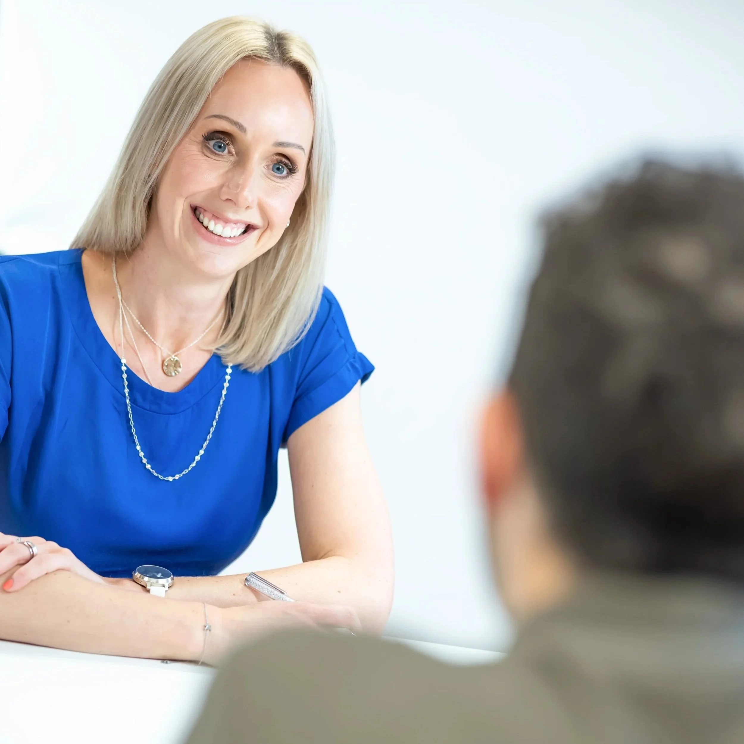 A woman with blonde hair smiling during a conversation with a man in the foreground, in a professional setting.