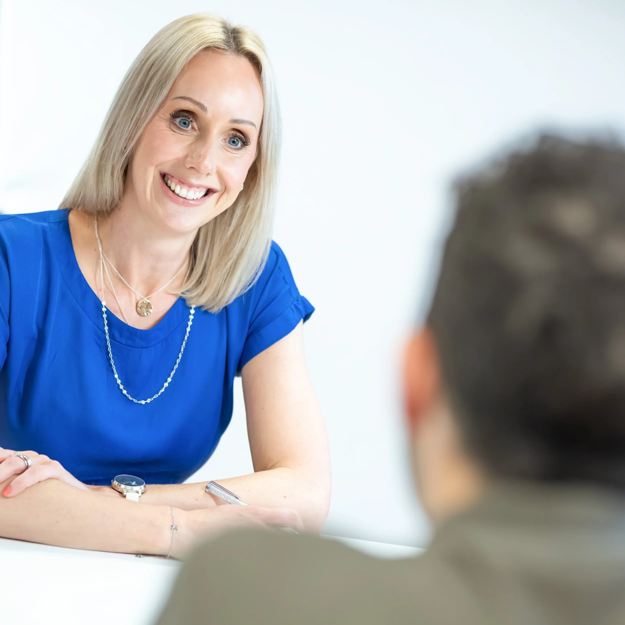 A woman with blonde hair and blue eyes smiling, sitting at a table and talking to a person whose face is not visible in the foreground, in a bright, clean setting.
