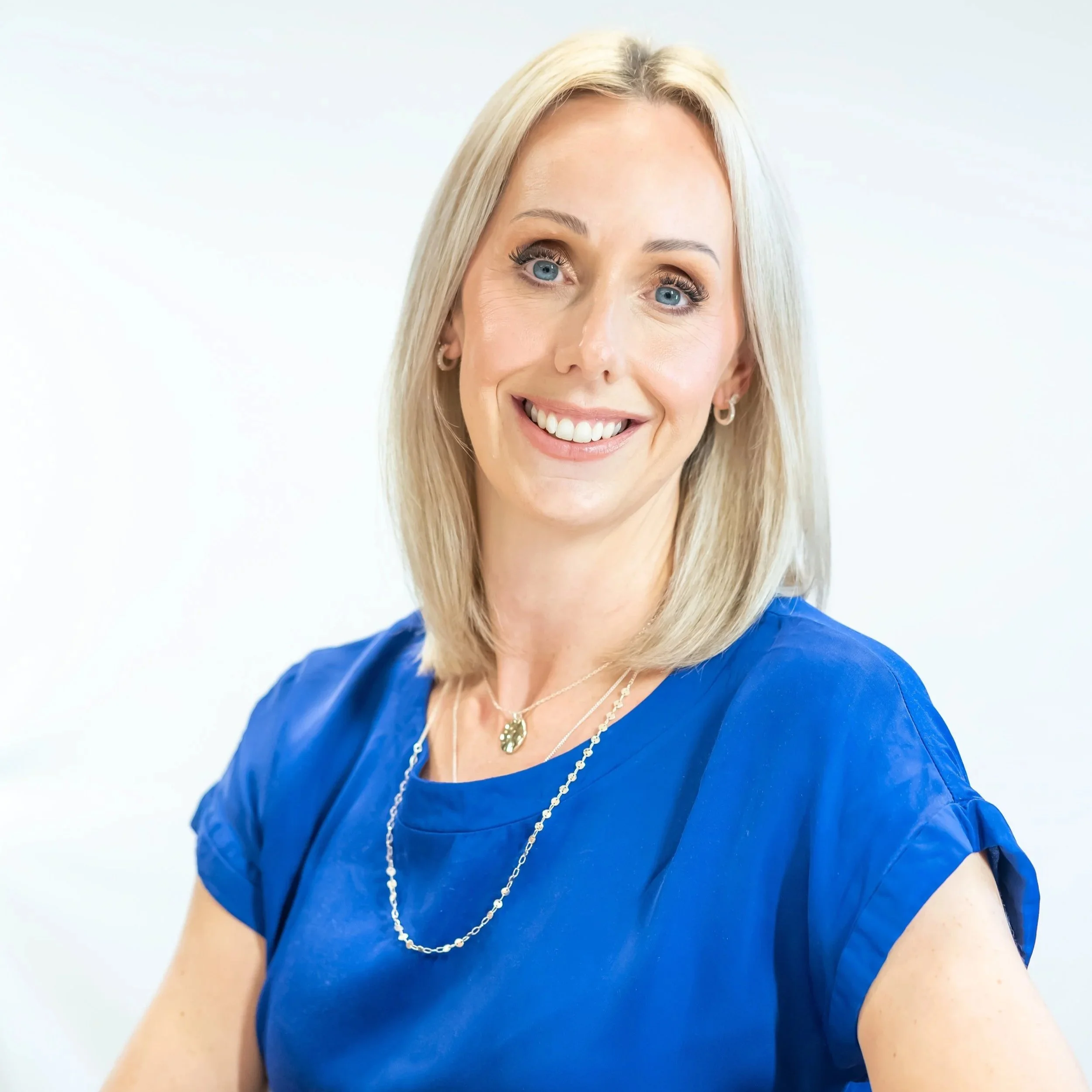 A woman with shoulder-length blonde hair, blue eyes, and light skin, smiling and wearing a blue blouse, gold earrings, and layered necklaces, with a plain white background.
