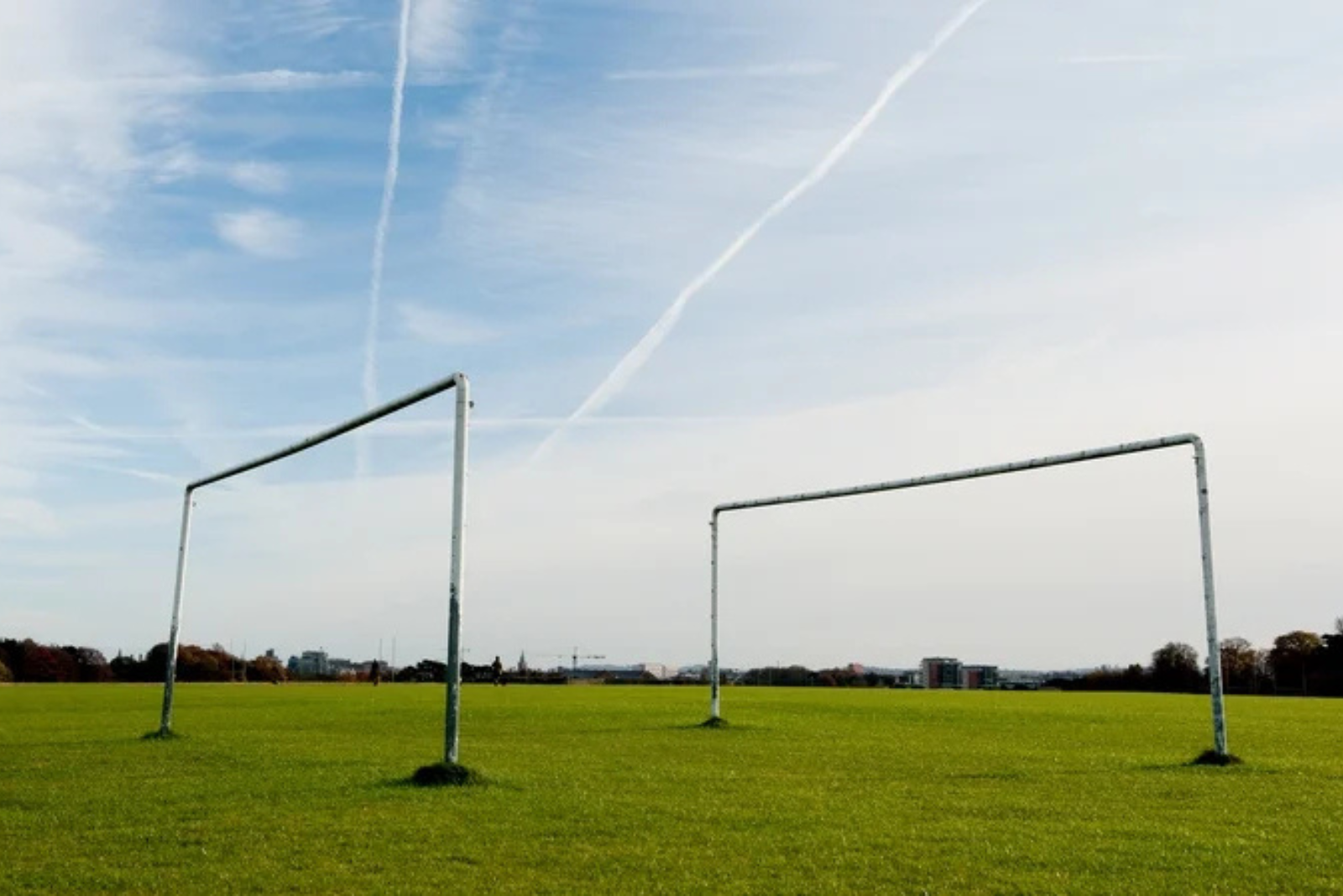 Two empty soccer goals on a grassy field under a blue sky with clouds and contrails.