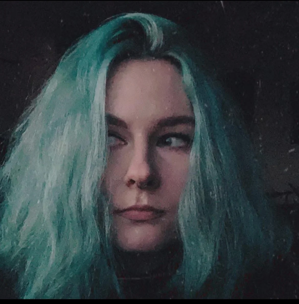 Close-up of a woman with long, wavy, teal-colored hair and light makeup, looking to the side with a neutral expression.