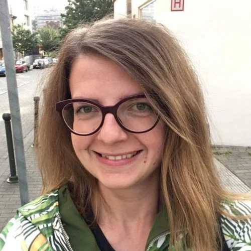 A woman with shoulder-length light brown hair, wearing glasses, smiling outdoors on a city street.