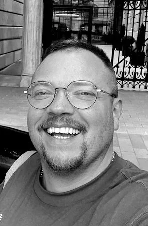 Lächelnder Mann mit Brille und geringem Bart, fotografiert im Freien mit einem Zaun und einer Hauswand im Hintergrund. Sebastian Reßler. Leiter und Designer im Estudio SR Designbüro. Mit Herz und Seele.