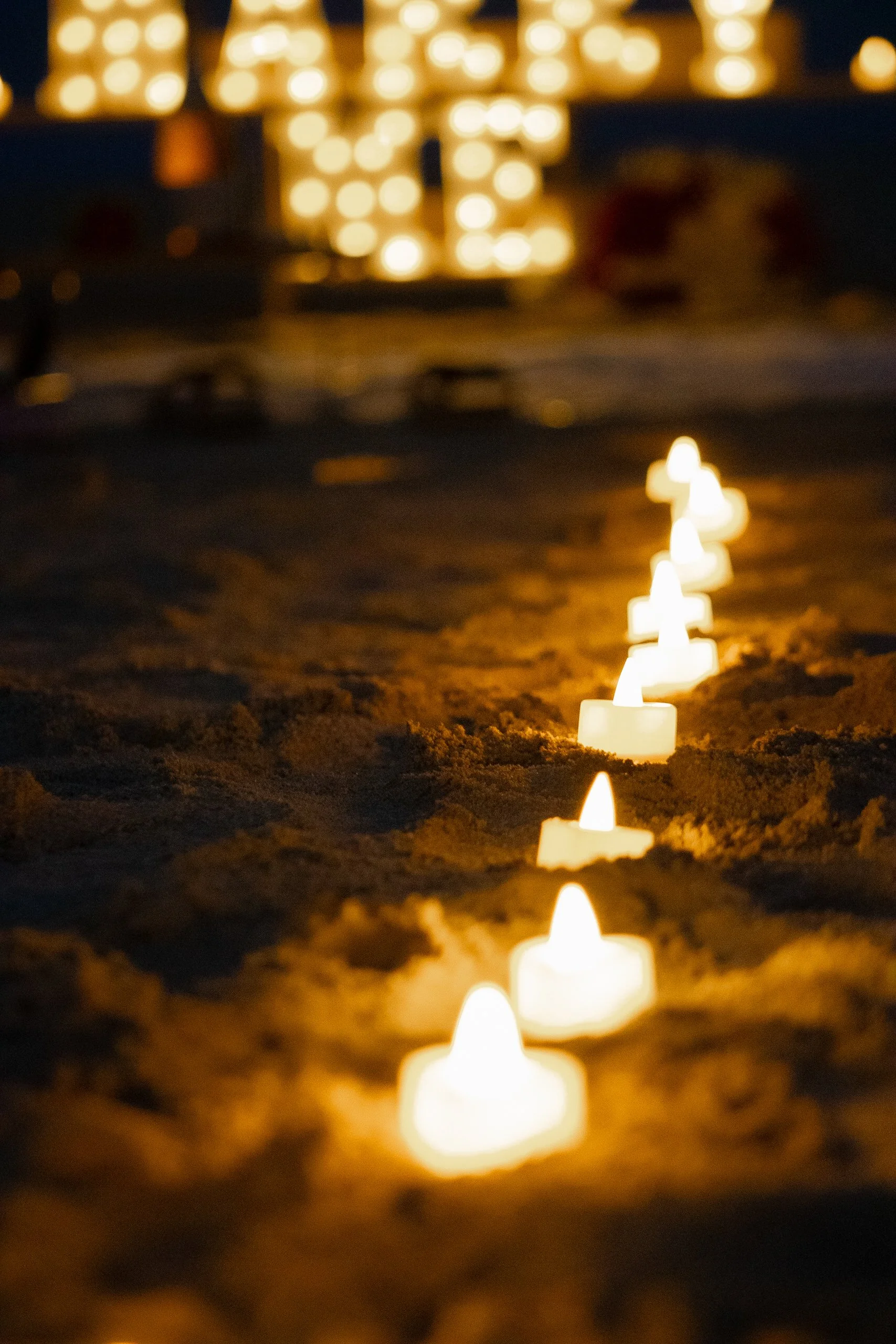 Row of small lit candles on sand at night with large blurred light display in the background.