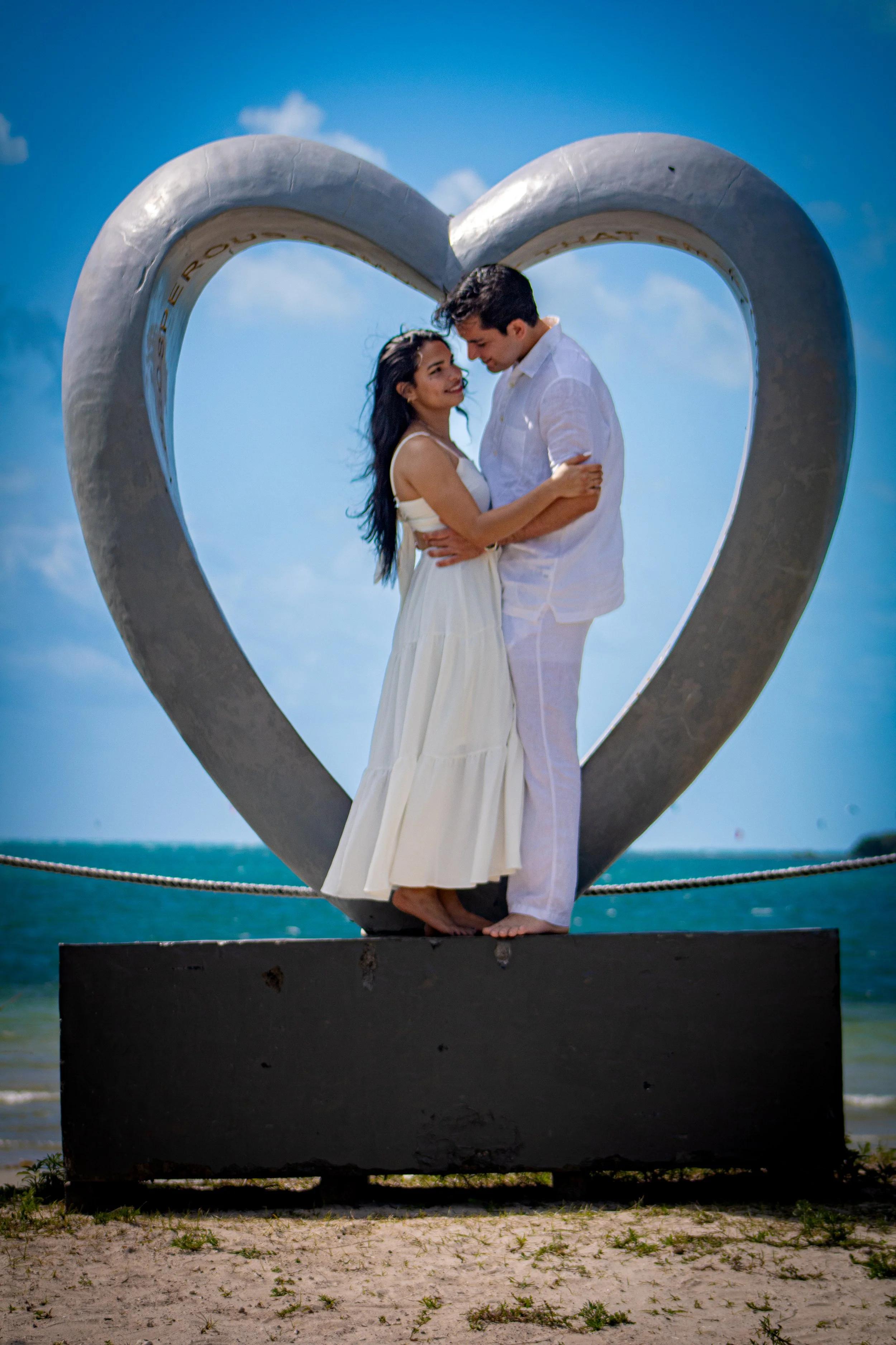 A couple standing close together inside a large heart-shaped sculpture on the beach, with the ocean and blue sky in the background.