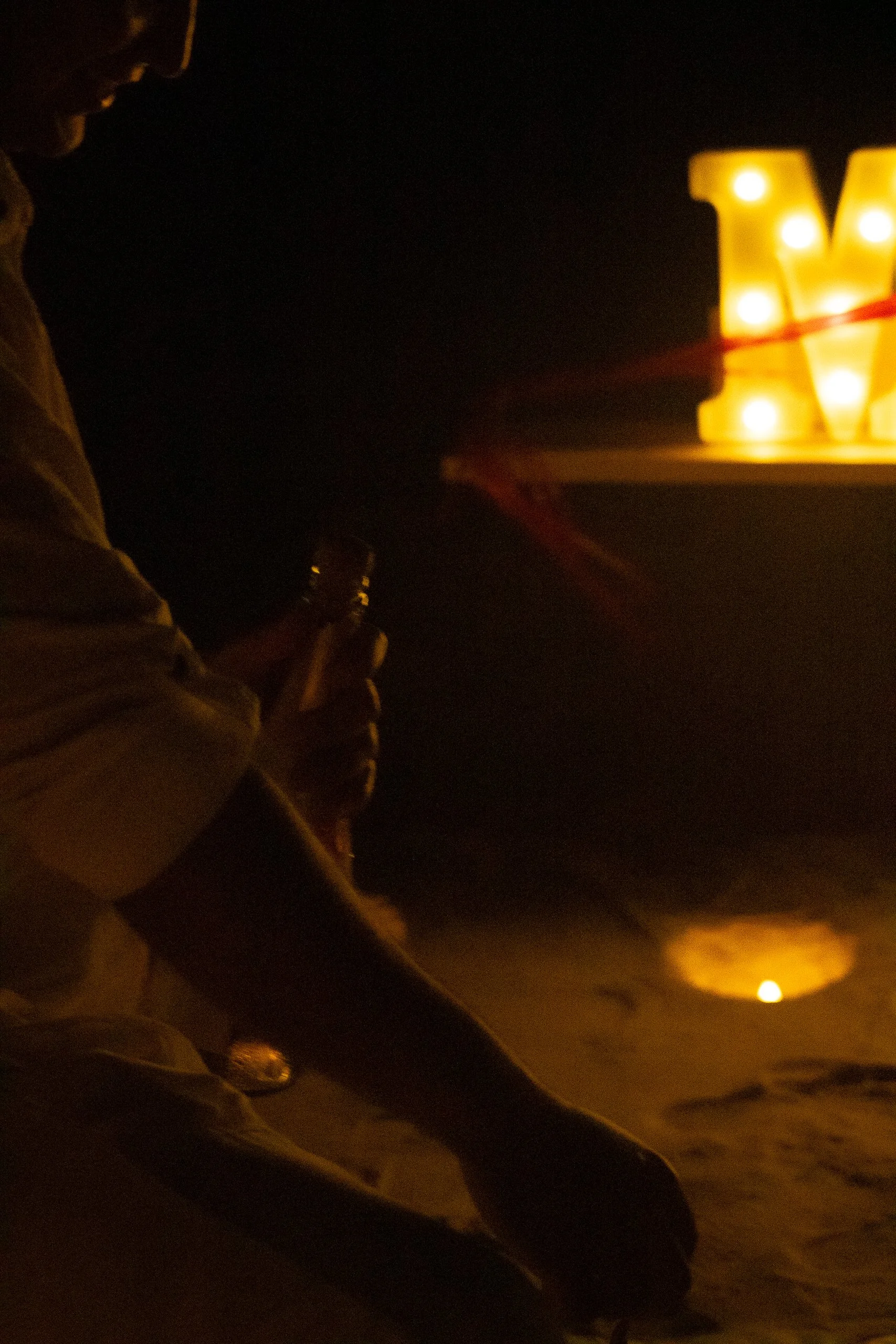 Silhouette of a person on the beach holding an object at sunset or sunrise, with a large illuminated letter 'M' in the background.