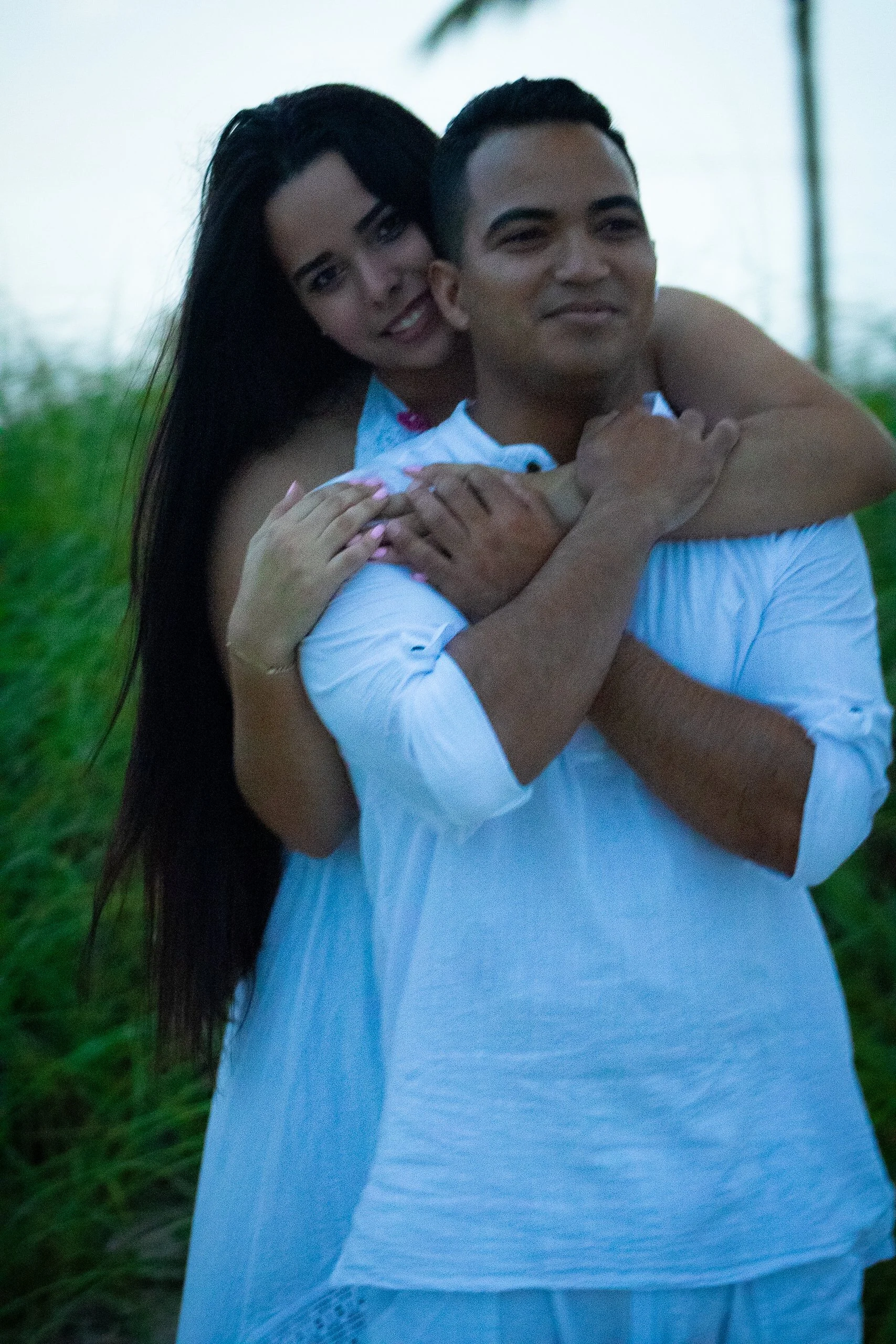 A couple standing outdoors in a grassy area during dusk, with the woman hugging the man from behind and both smiling softly.
