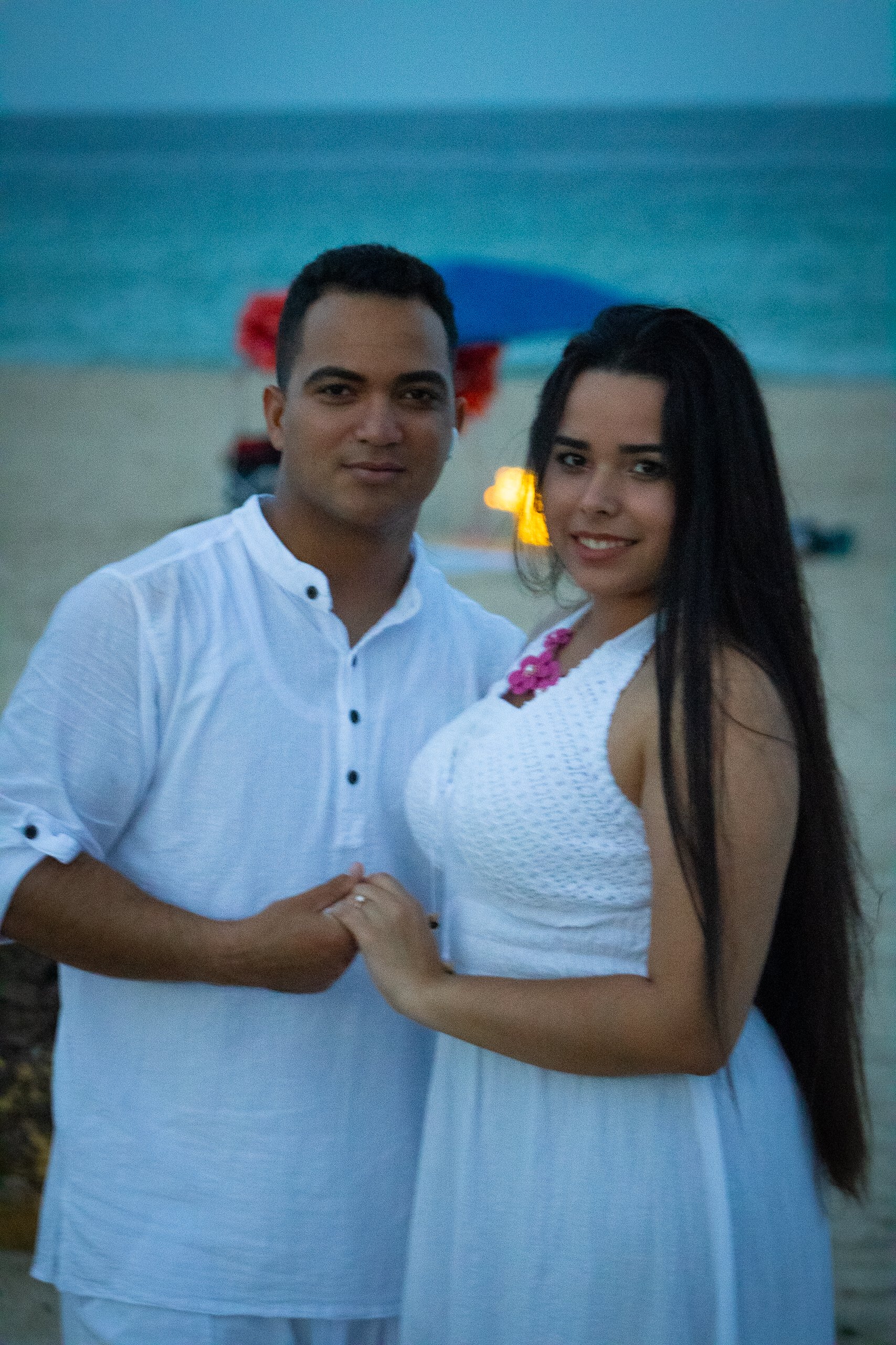 A young couple holding hands on a beach at dusk, with the ocean in the background and a colorful umbrella nearby.