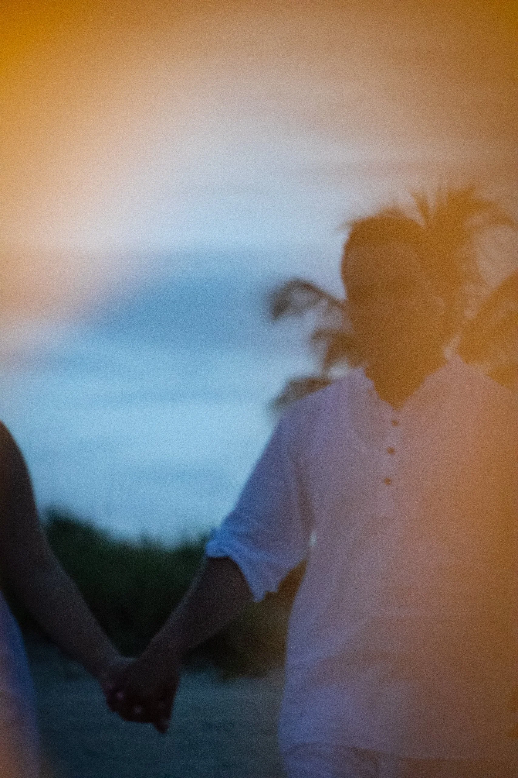 A man holding hands with someone during sunset, with a blurred background of palm trees and sky.