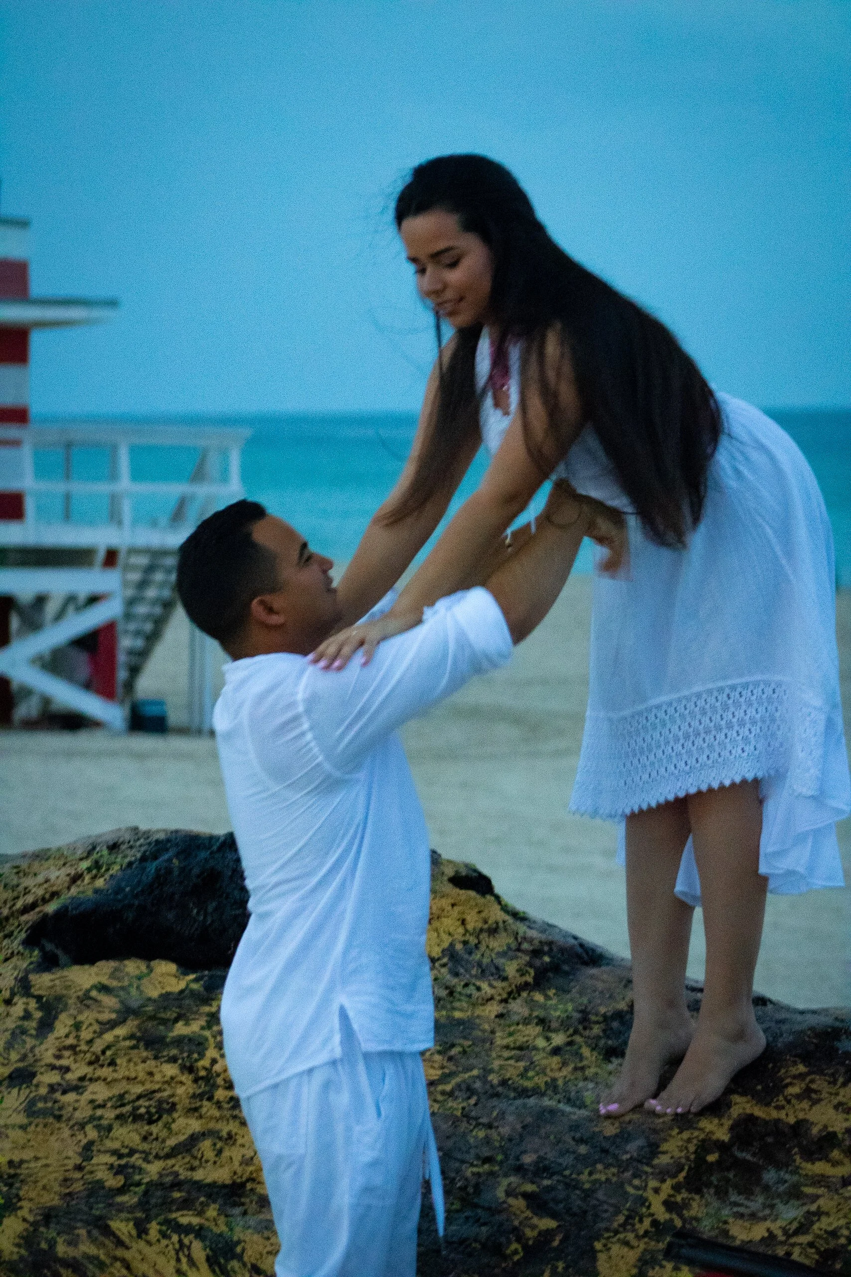 A couple on the beach, with the man holding the woman up, both dressed in white, during sunset or dusk.