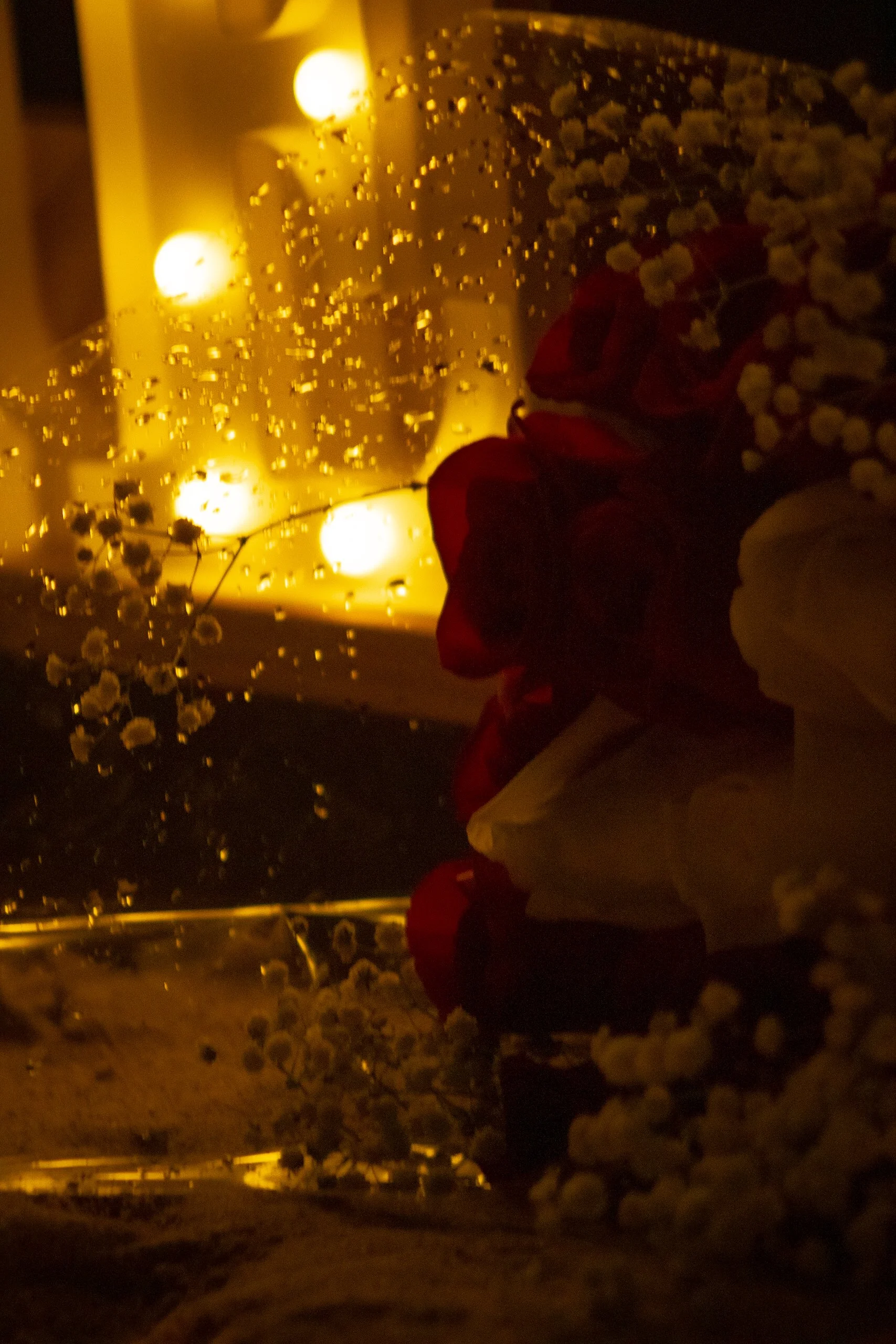 A view through a rain-covered window with raindrops on the glass, showing a reflection of a person dressed warmly, standing amongst flowers, at night with warm yellow lighting.