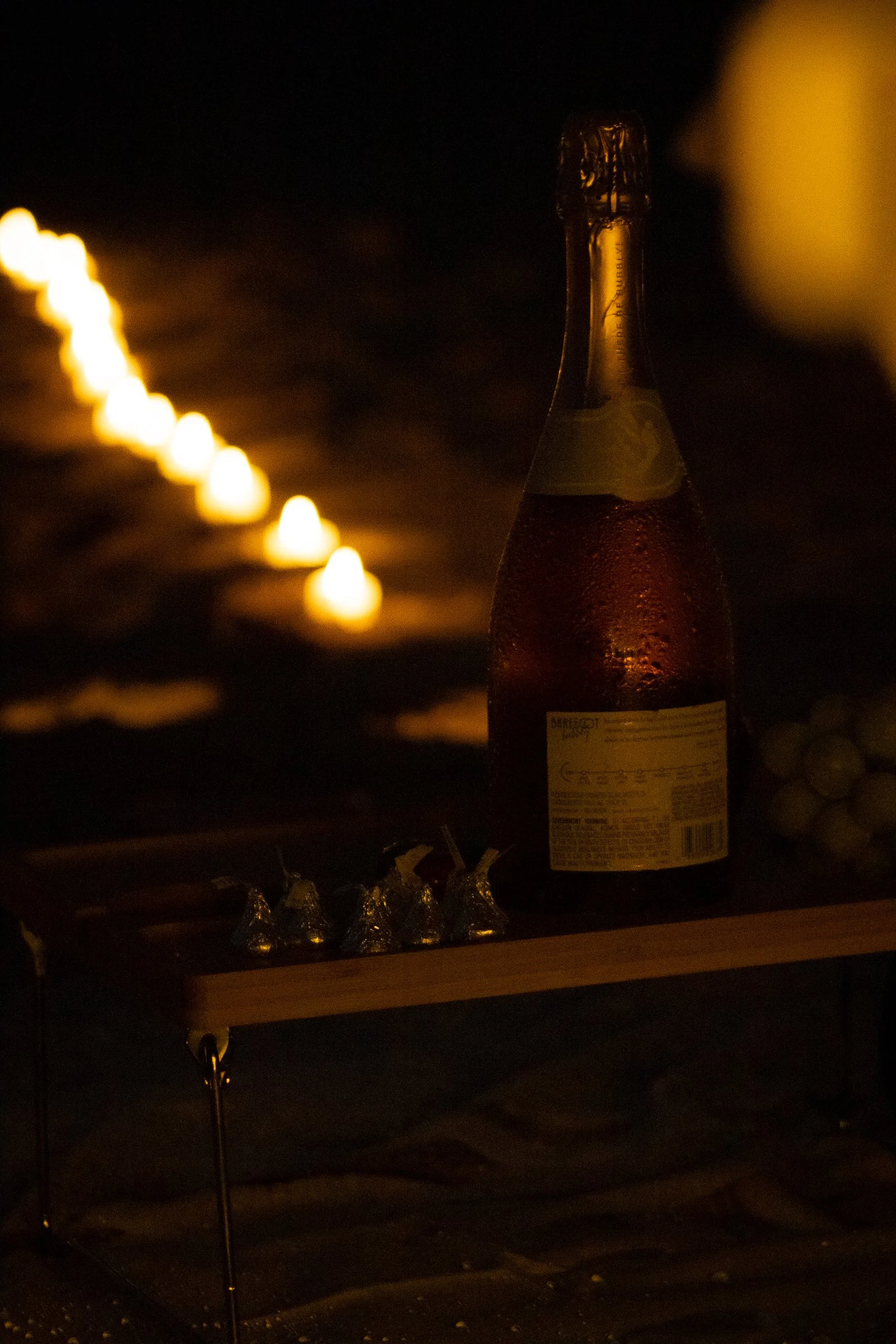 A bottle of champagne on a table with small chocolates wrapped in foil nearby, in a dimly lit setting with a row of candles in the background.