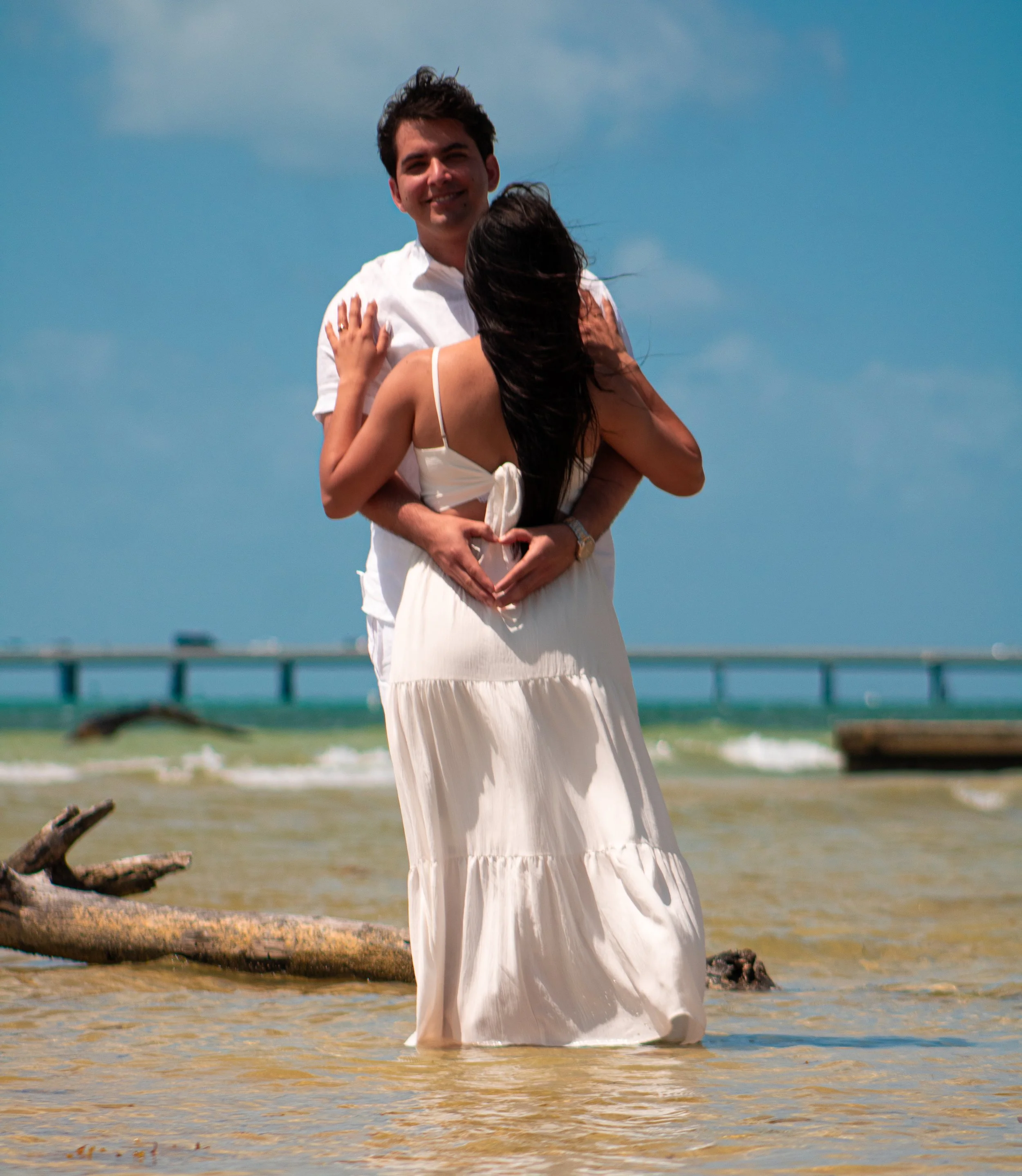 A couple embracing on a beach, with the man smiling and the woman with long dark hair, forming a heart shape with their hands over her stomach.