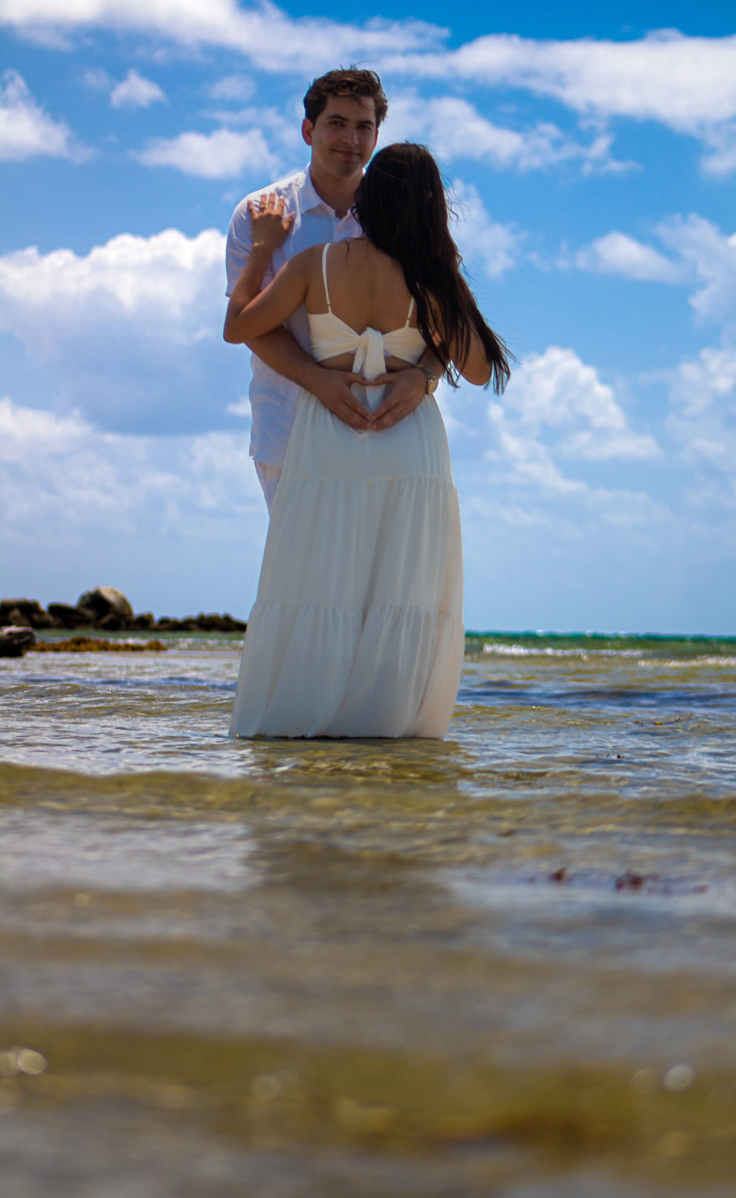 A couple standing in shallow ocean water on a sunny day, with the woman forming a heart shape with her hands on her belly and the man standing behind her, both facing the camera.