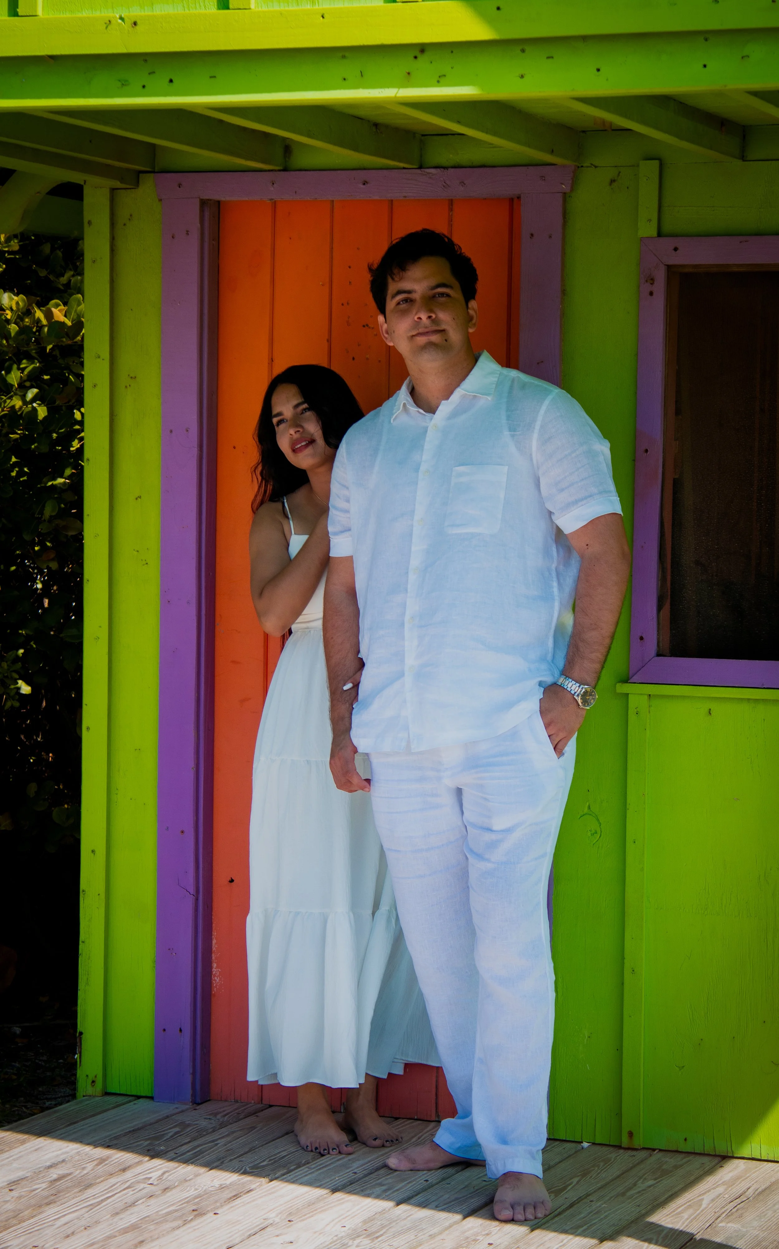 A man and woman dressed in white standing outside a colorful wooden structure with purple, orange, and green panels, on a sunny day.