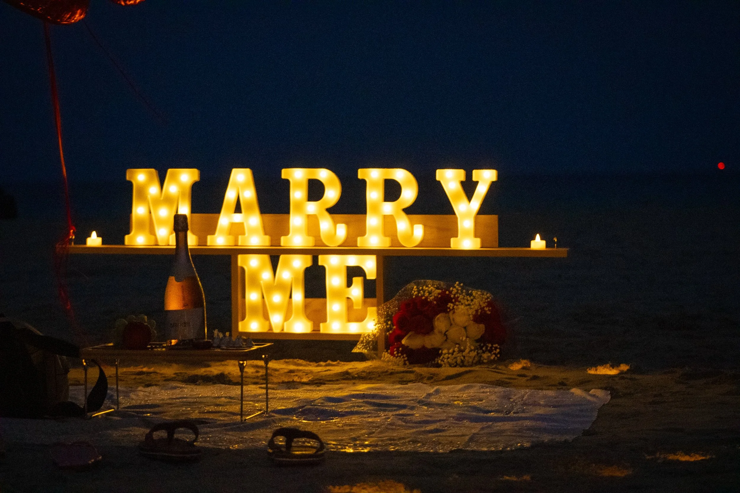 Large illuminated marquee letters spell out "MARRY ME" beside a bouquet of flowers, a bottle of champagne, and candles on a beach at night.