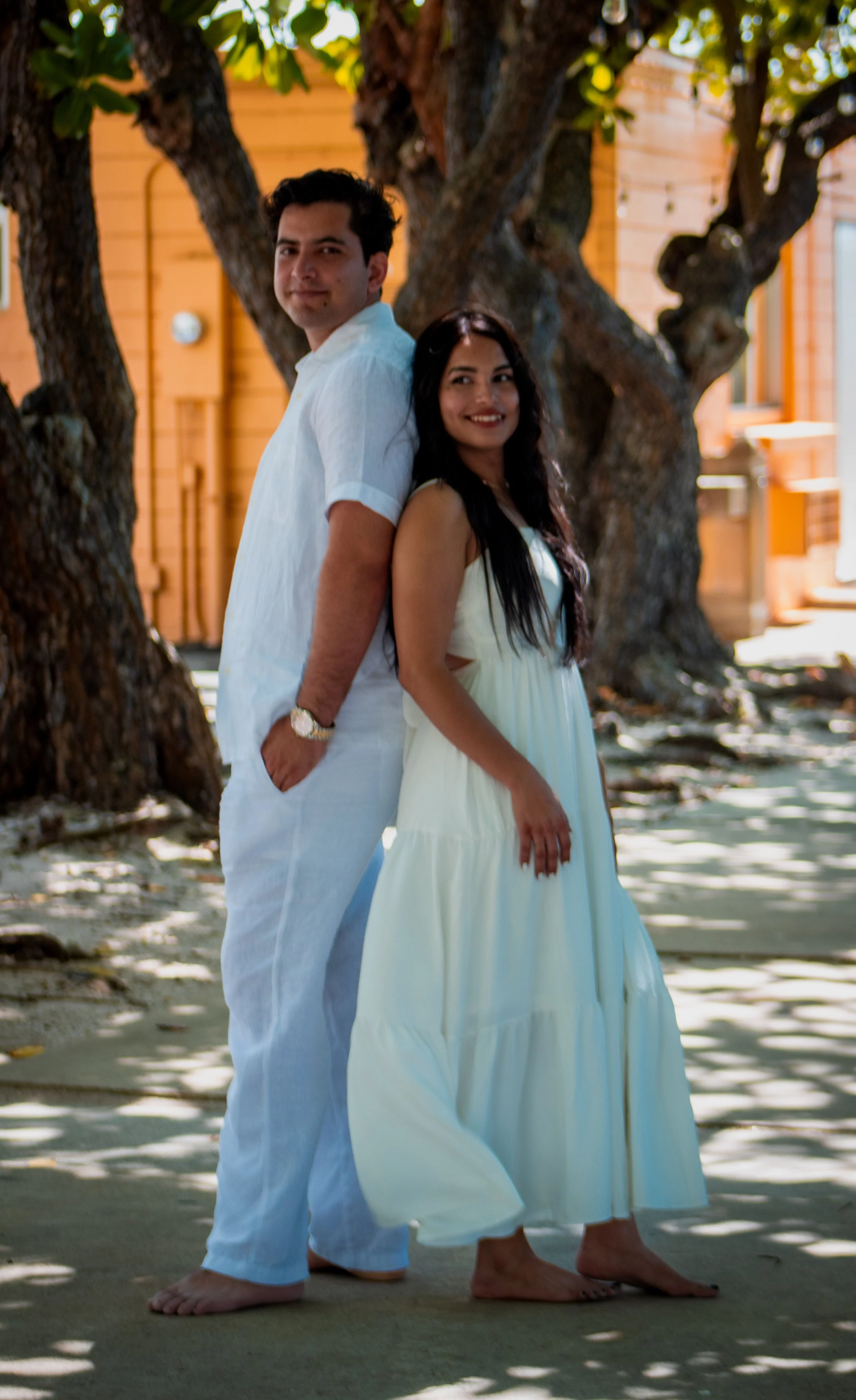 A couple in white standing outdoors, back to back, under a large tree with a wooden building in the background.