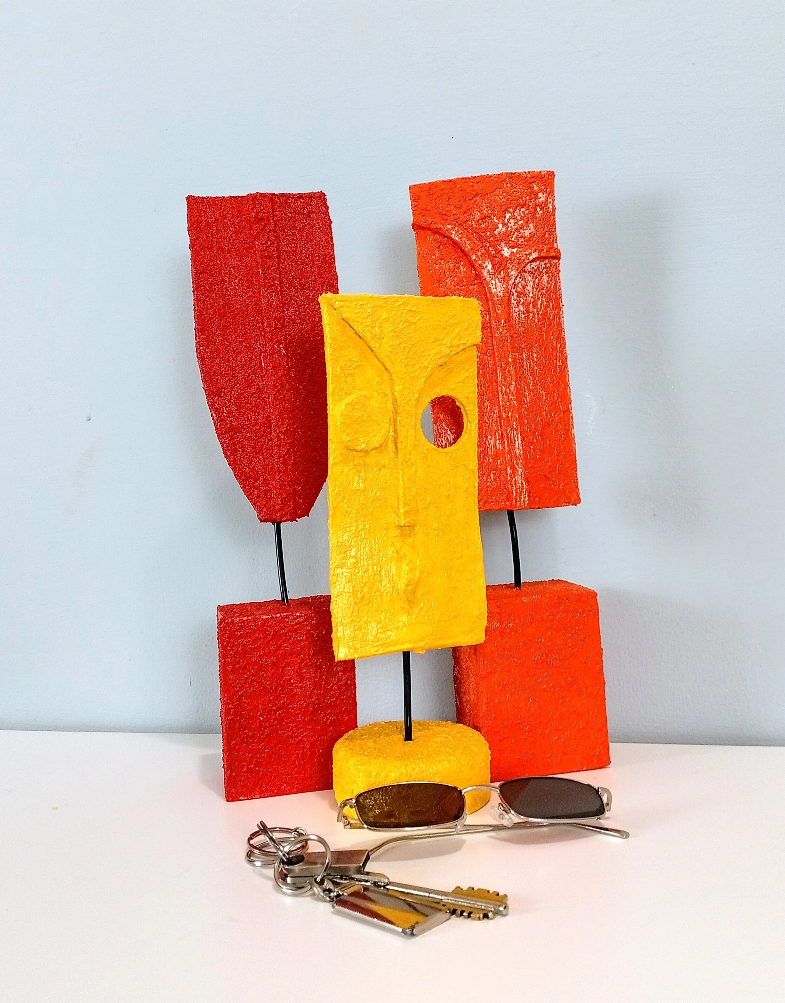 Yellow – Minimalist Abstract Figurative Sculpture with red and orange sculptures in the background, size reference with sunglasses and keys.