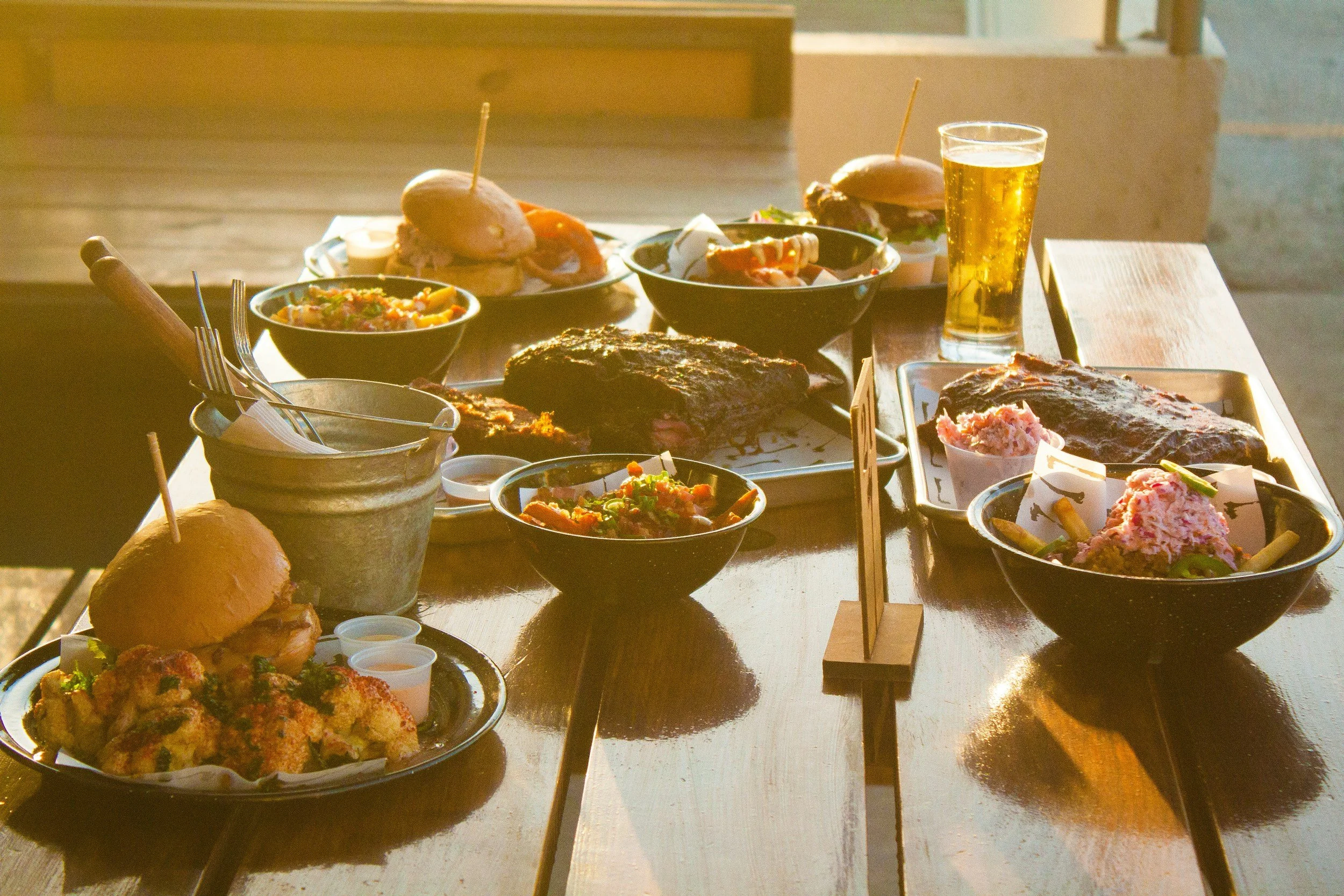A variety of grilled and plated foods on a wooden table, including burgers, bowls of salad or side dishes, and a glass of beer in a bright setting.